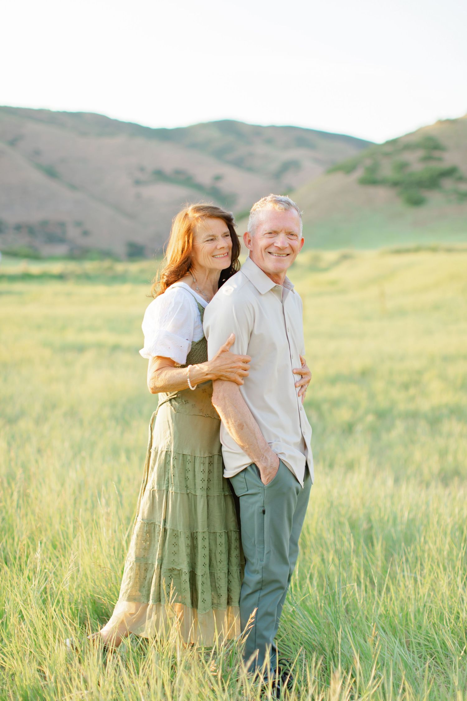 A couple shares a tender moment while walking through a green meadow with rolling hills in the background.