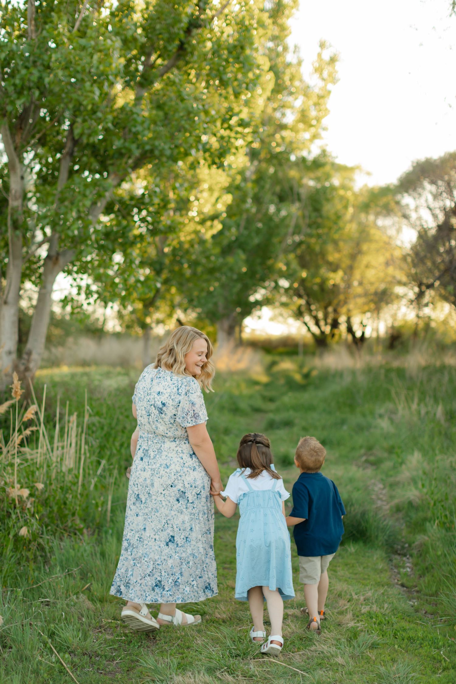 Family walking together on a grassy path surrounded by green trees during golden hour sunset.