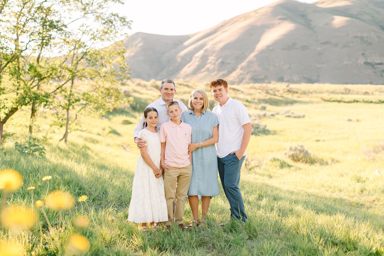 Family of five posing together in a sunny green meadow with rolling hills in the background during golden hour.