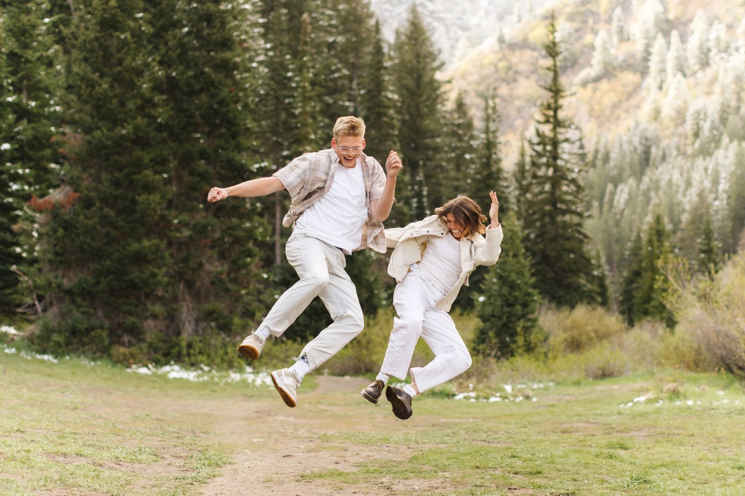Joyful couple jumping together in matching white outfits on a mountain trail surrounded by pine trees.