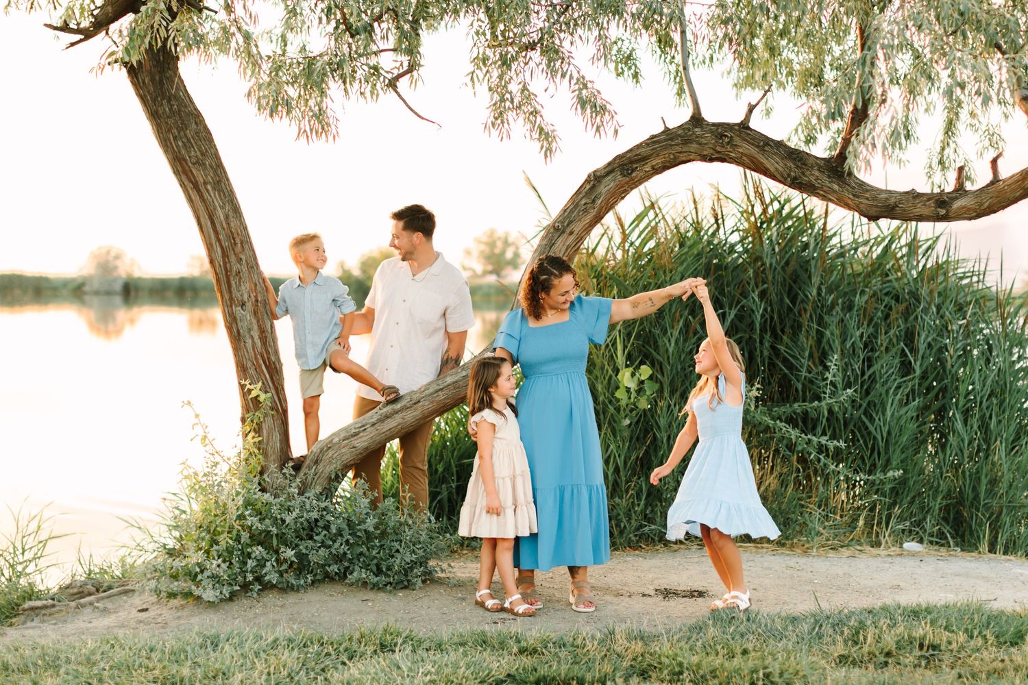 Happy family of five enjoying a sunny day by the lake, with mother twirling daughter near a tree in Bountiful, Utah.