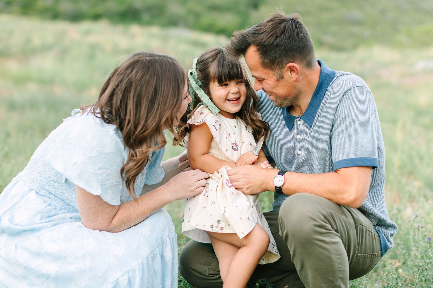 Happy family moment with parents lovingly embracing their laughing toddler daughter outdoors in a green meadow.