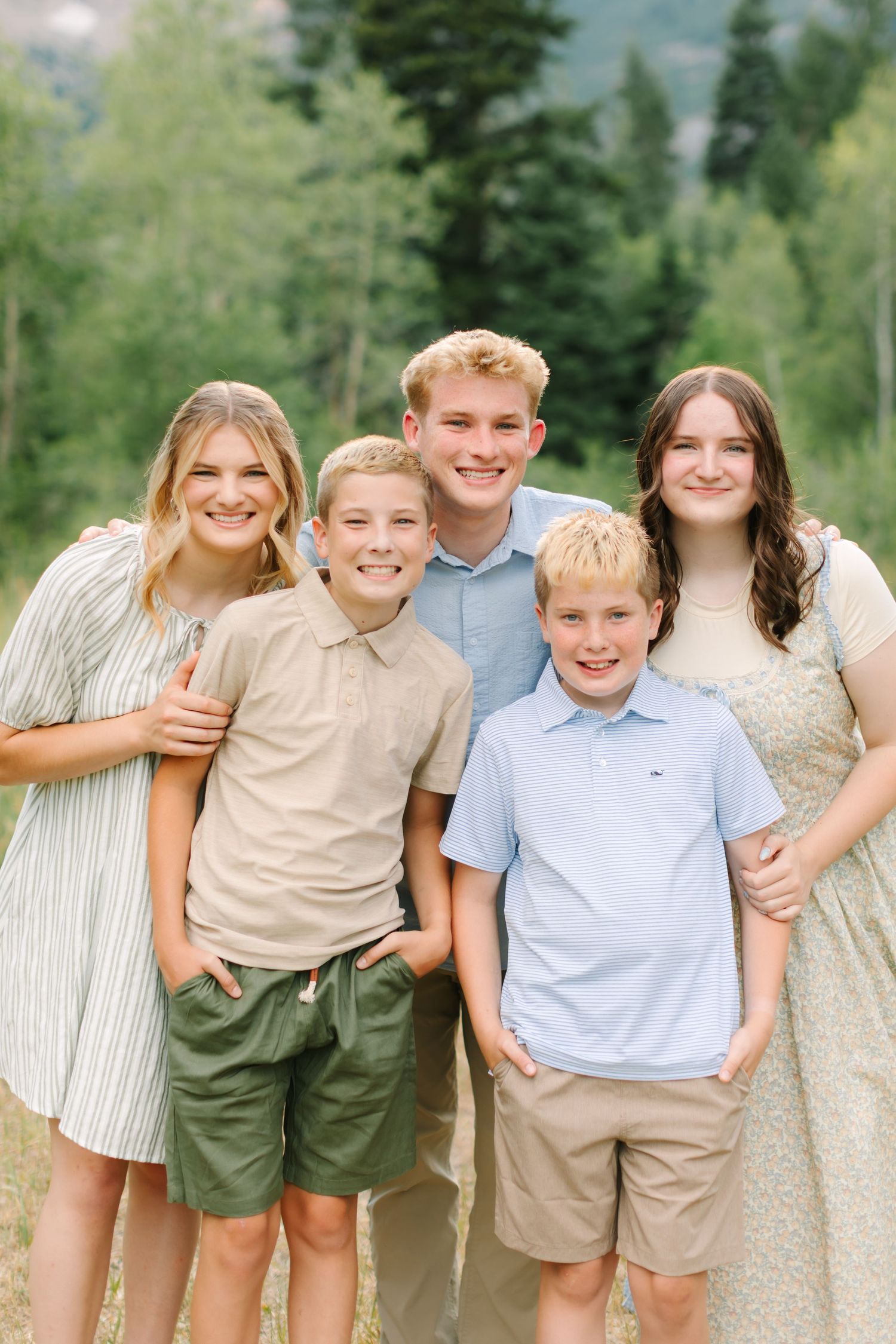 Five smiling children pose together outdoors in a lush green mountain setting wearing casual summer outfits.