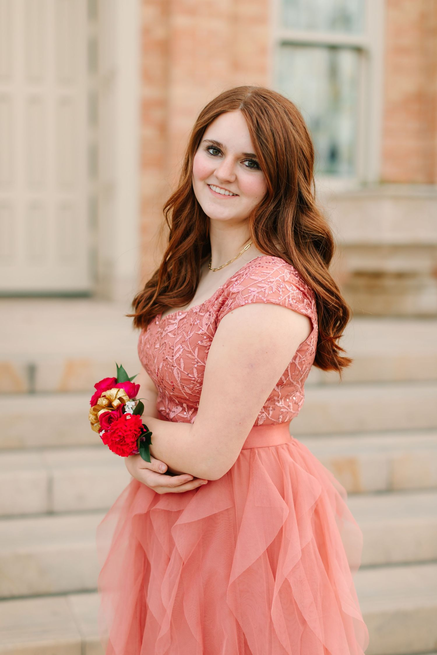 A person in a coral lace dress with tulle skirt poses outdoors holding red flowers against a brick building.