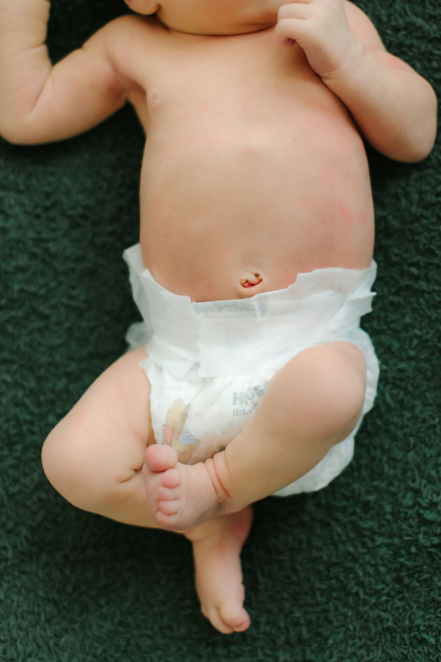 Baby wearing white diaper lying on dark green carpet showing tiny feet and legs.