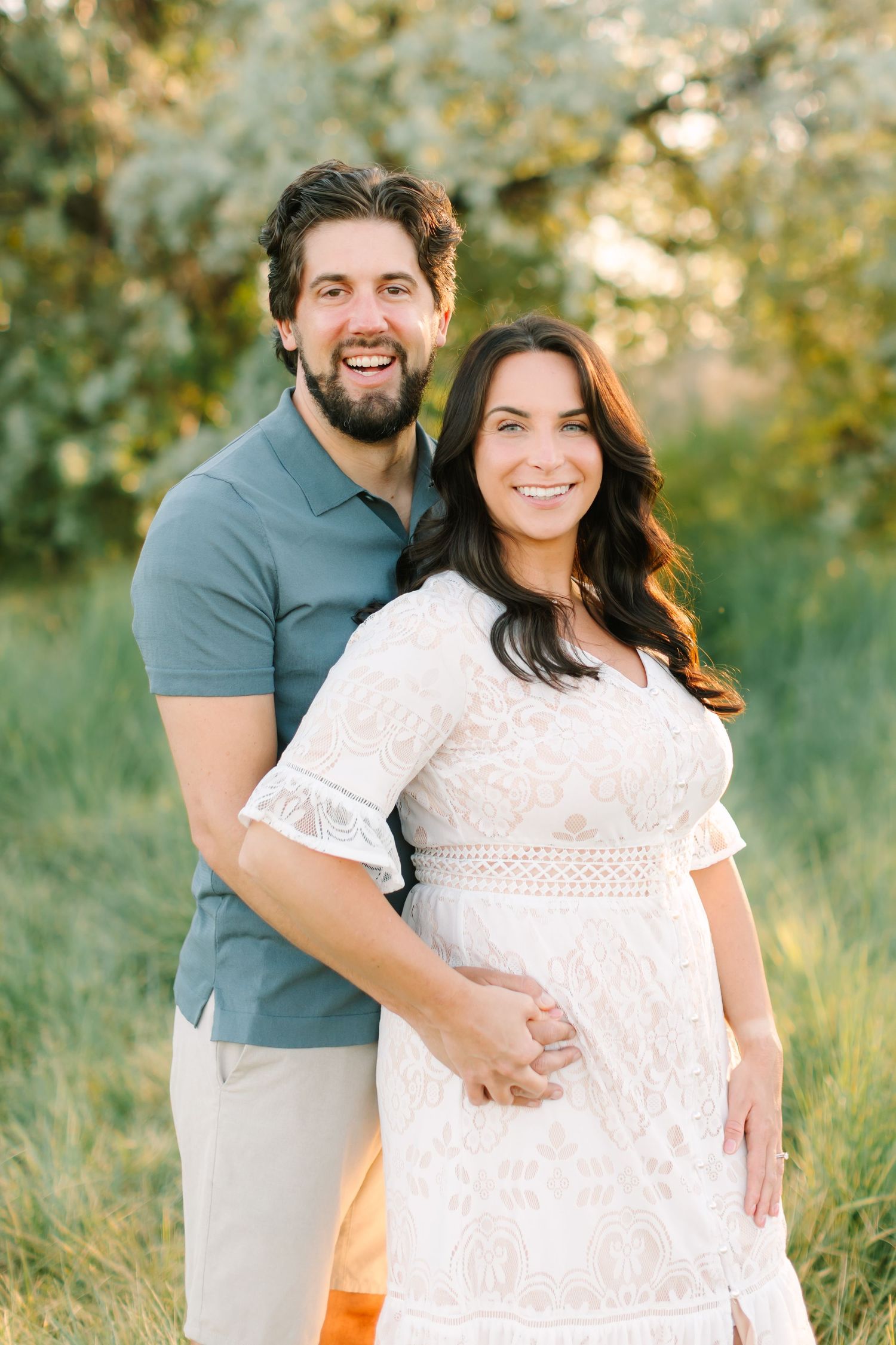 Smiling couple posing outdoors, pregnant woman in white lace dress, man in blue polo, green trees background.