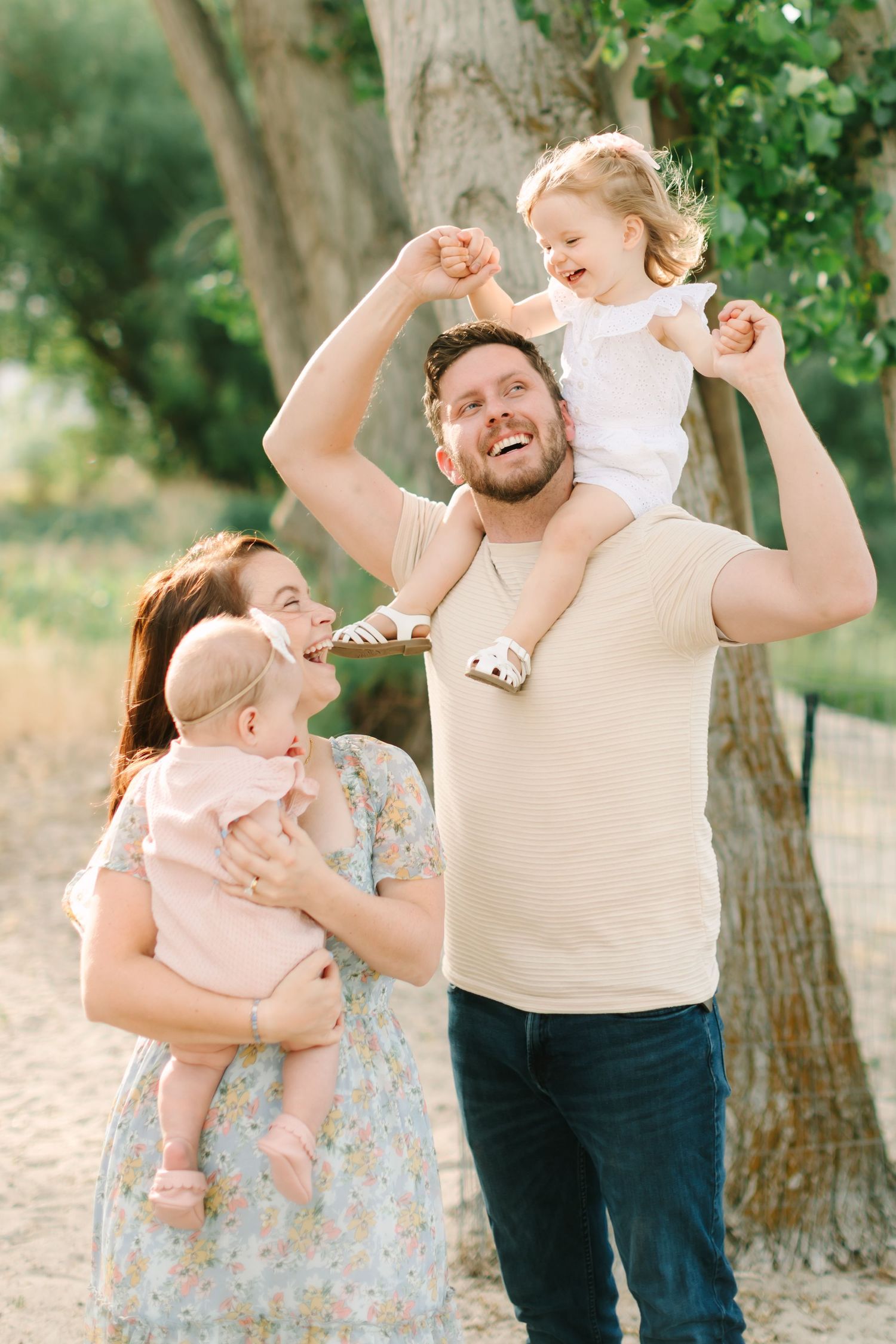 Happy family outdoors, parents holding two young children near trees in warm sunlight, smiling and laughing together.