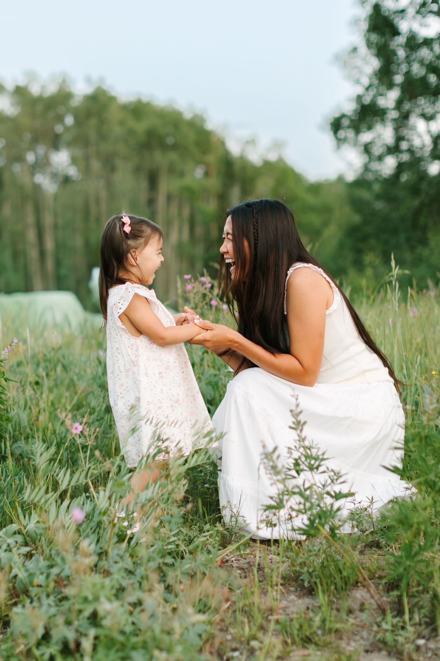 A mother and young daughter in white dresses share a joyful moment holding hands in a lush green field.