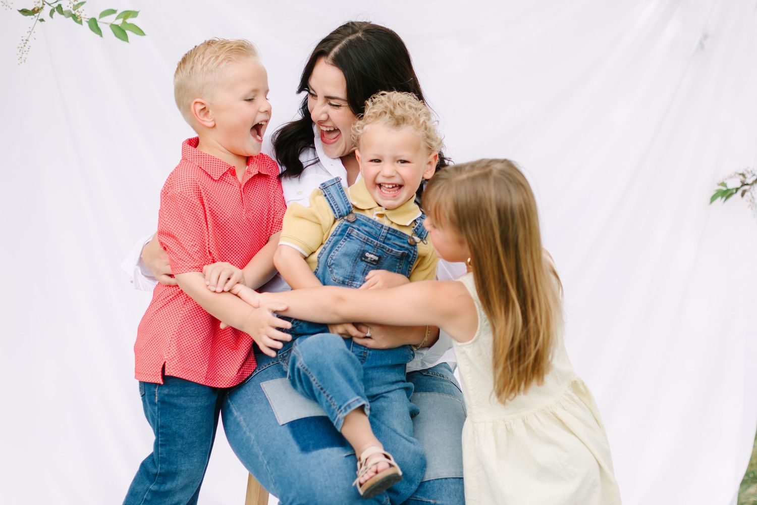 A joyful mother holds a laughing toddler in overalls while two siblings play together against a white backdrop for their motherhood photos,