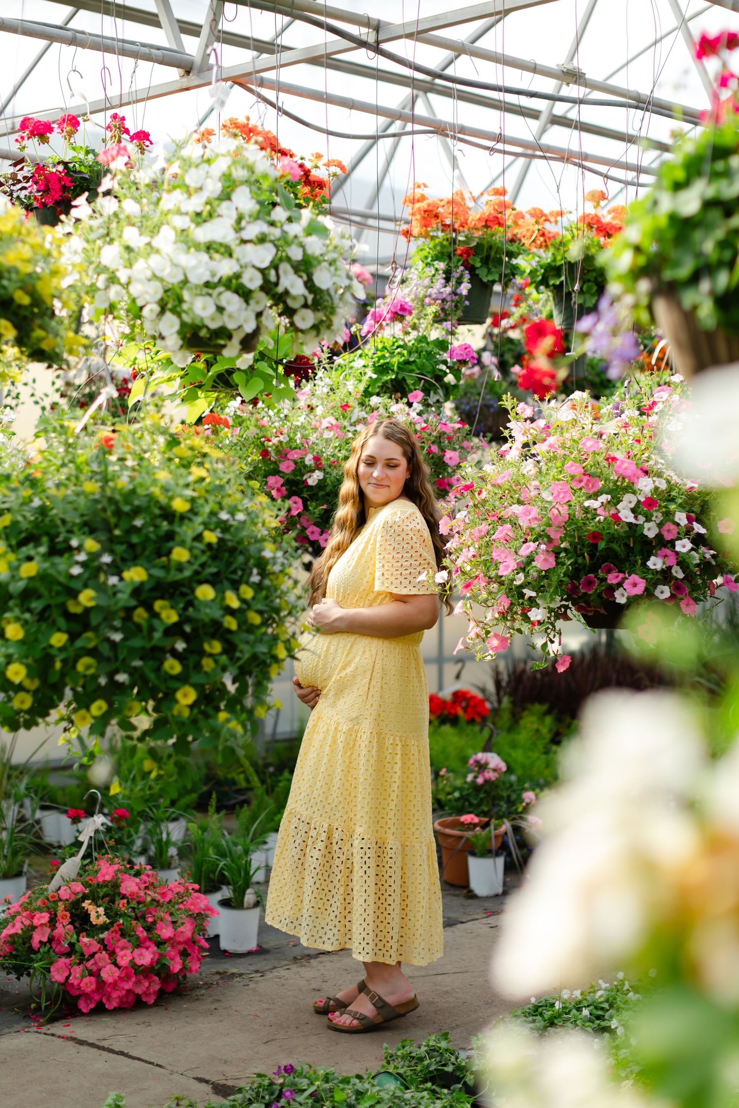 Yellow polka dot dress photographed in a greenhouse filled with colorful hanging flower baskets and blooming plants.