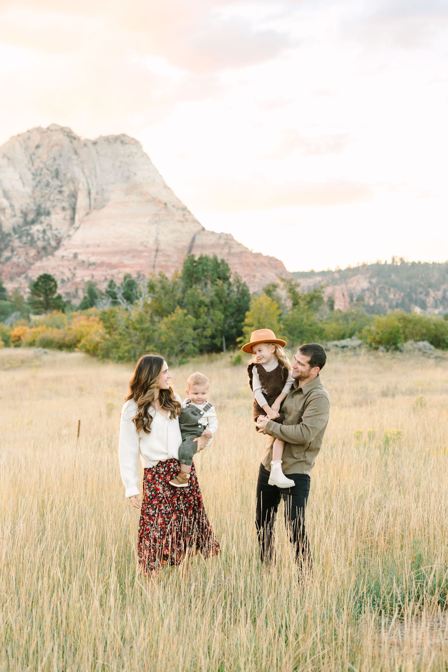 A family stands together in a golden field with red rock mountains and autumn foliage in the background.