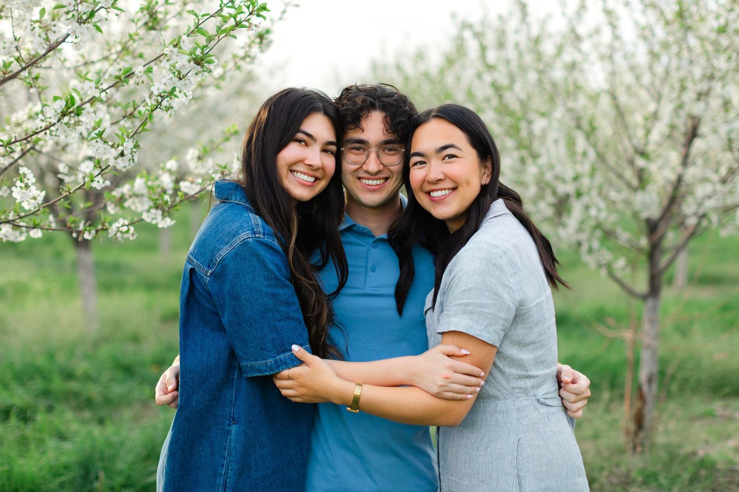 Three friends embrace while smiling together in a blooming spring orchard with white flowers.