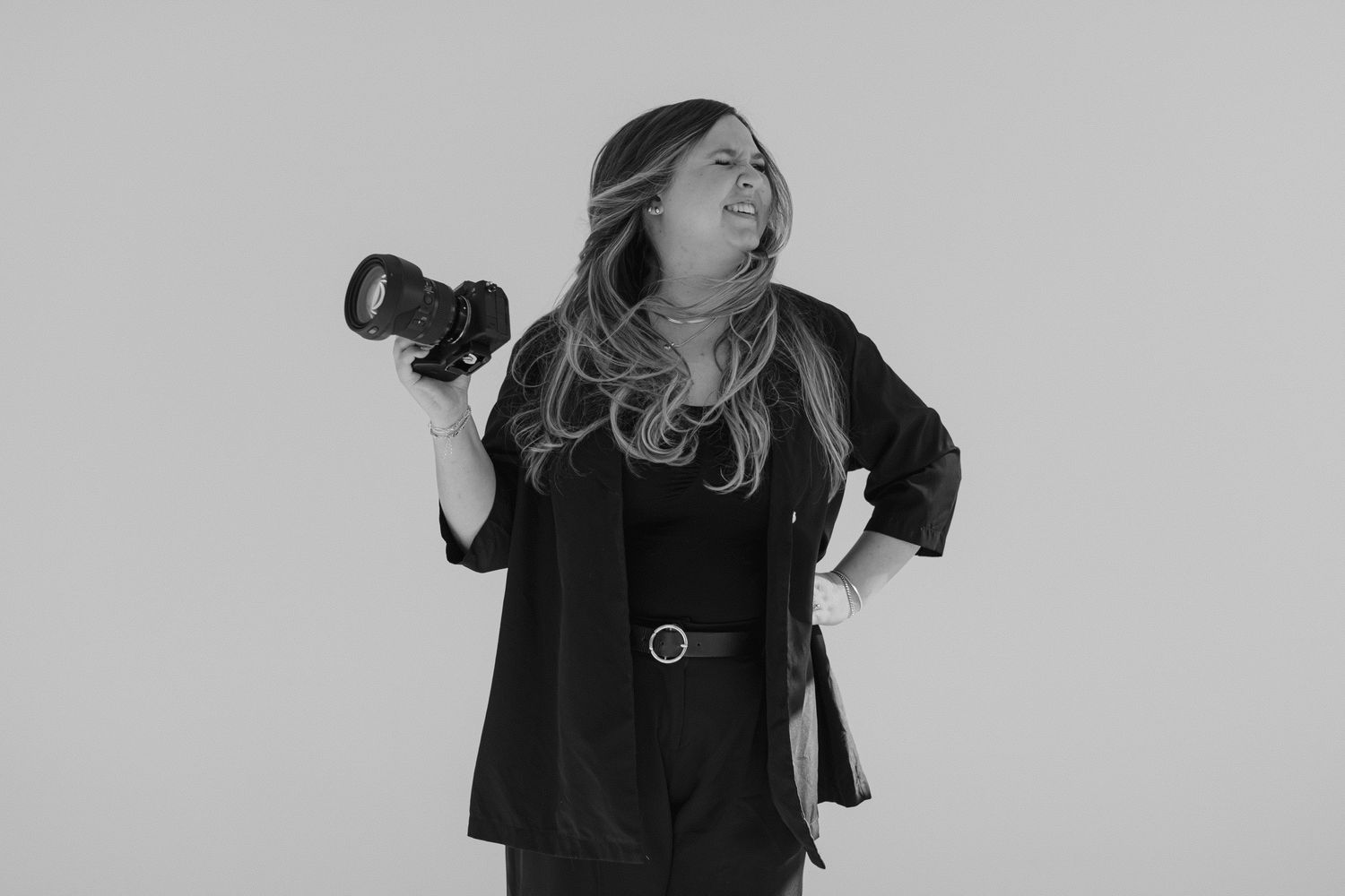 Professional photographer wearing black clothing holds a camera while posing against a white background.