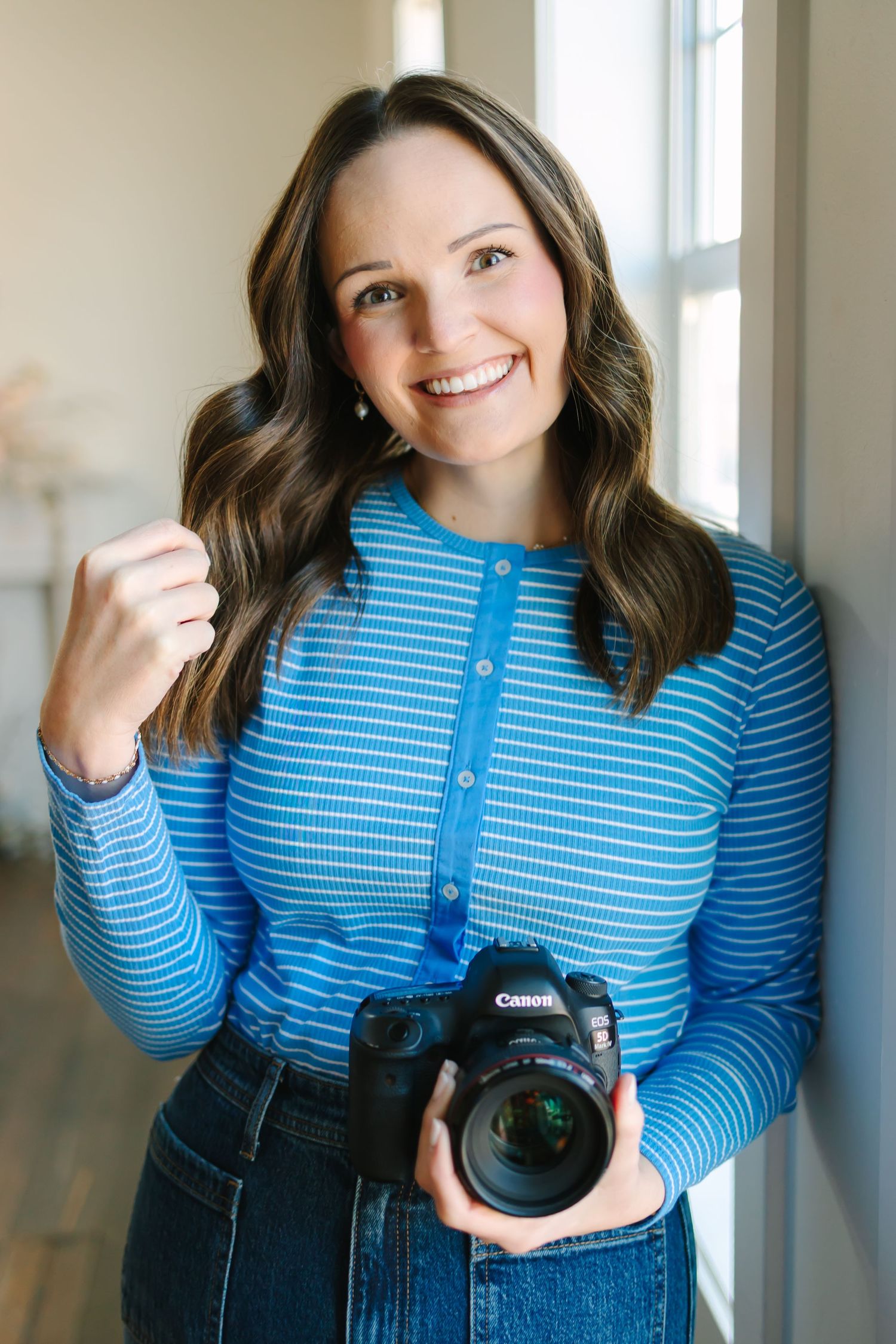 A photographer wearing a blue striped top holds a professional camera while posing against a bright window background in Utah County
