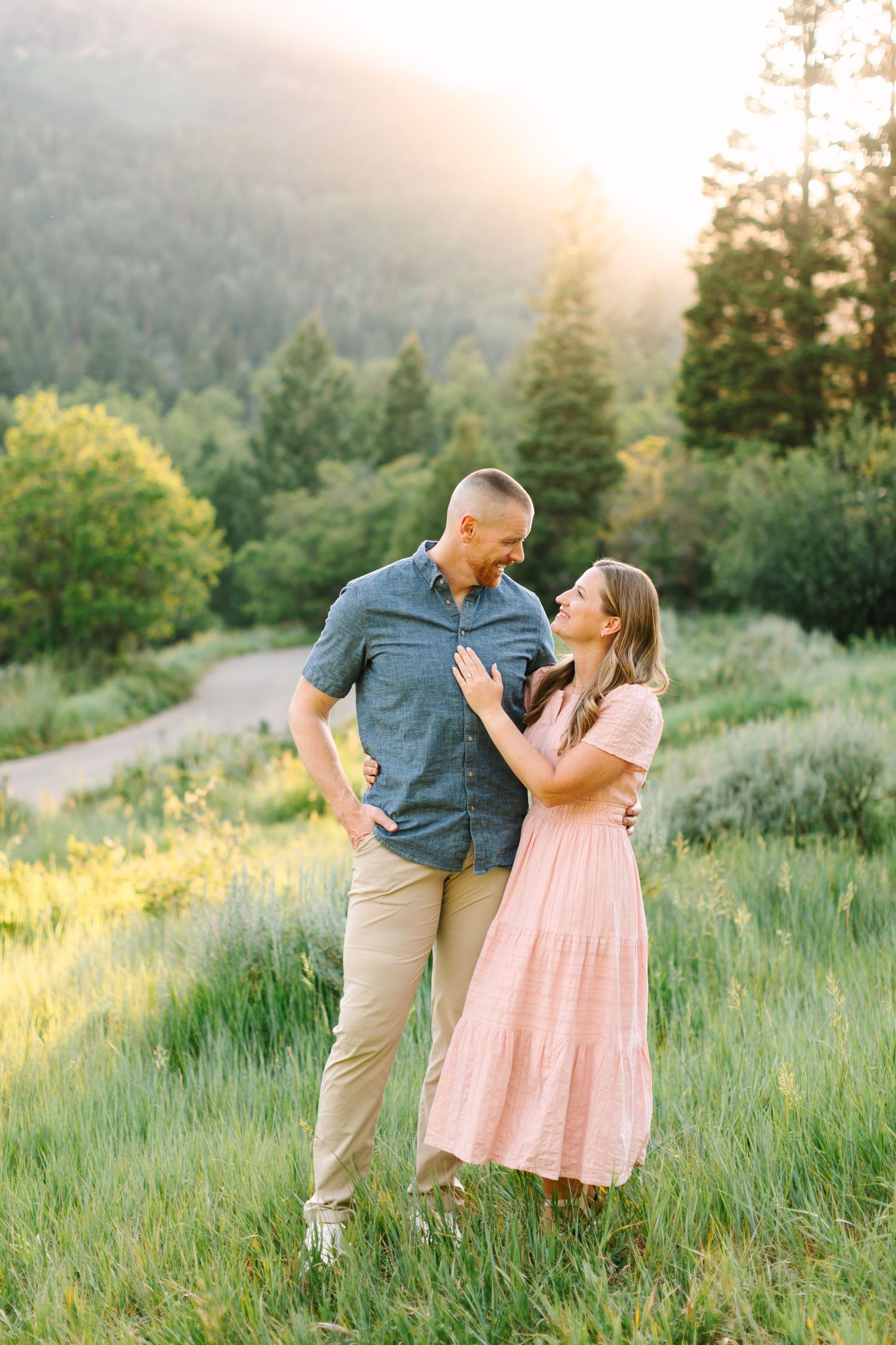 Couple sharing a tender moment in a sunlit meadow, man in blue shirt and woman in pink maxi dress, with forest backdrop.