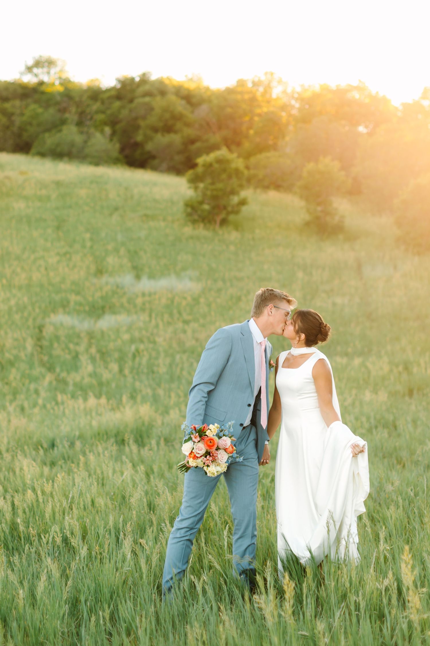 Bride and groom share a romantic kiss in a sunlit green field, groom in light blue suit holding a colorful bouquet.