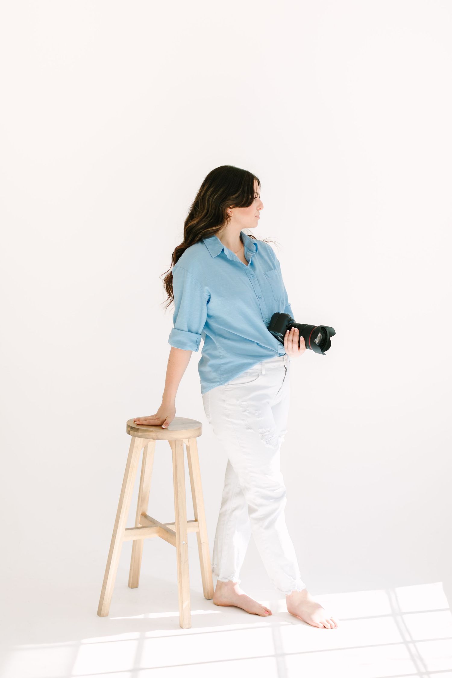 A photographer in casual attire sits on a wooden stool while holding a camera in a white studio.