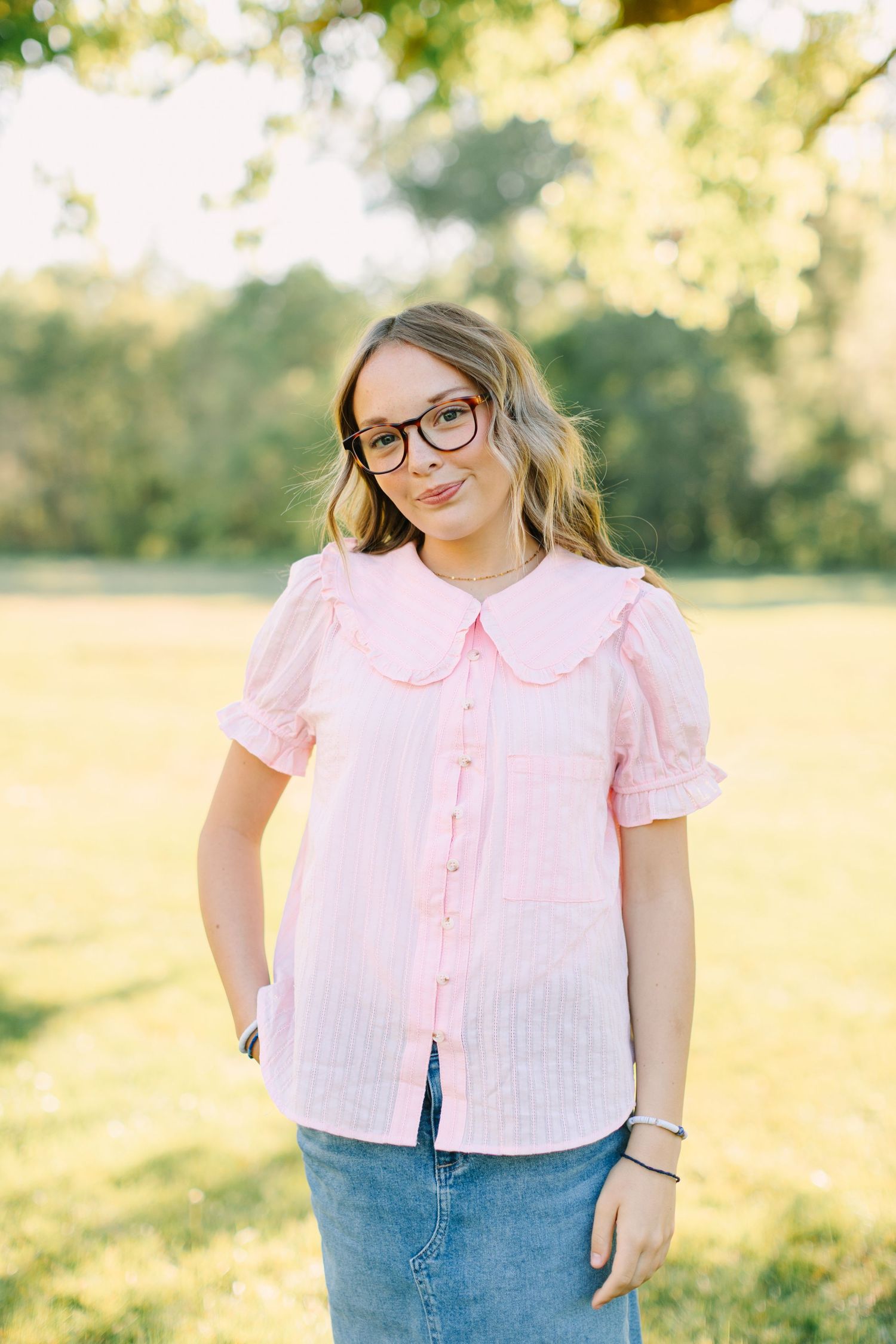 A person wearing glasses and a pink peter pan collar blouse poses outdoors in a sunny field.