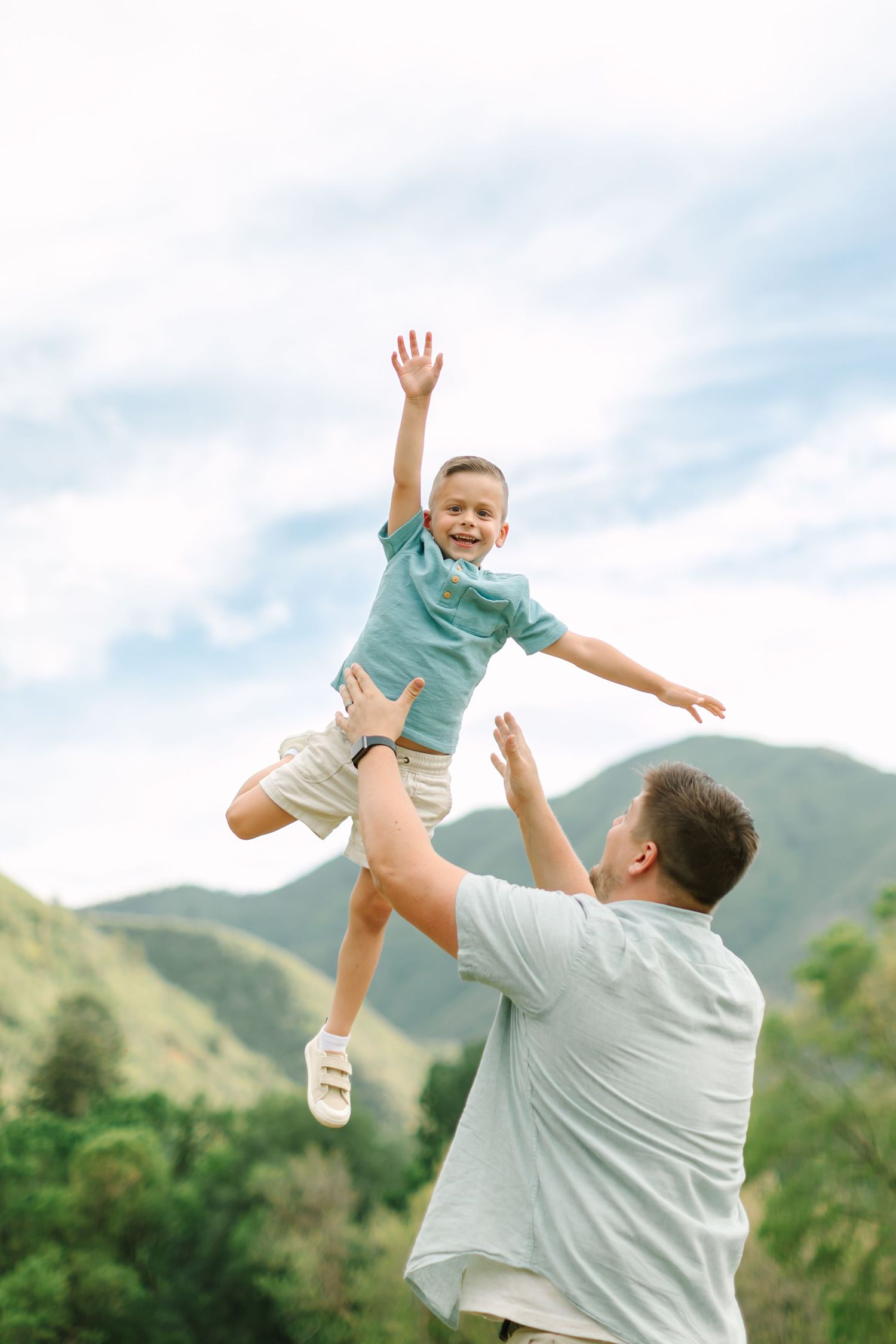 A parent playfully lifts a child wearing a teal shirt into the air against a backdrop of green mountains and cloudy sky.