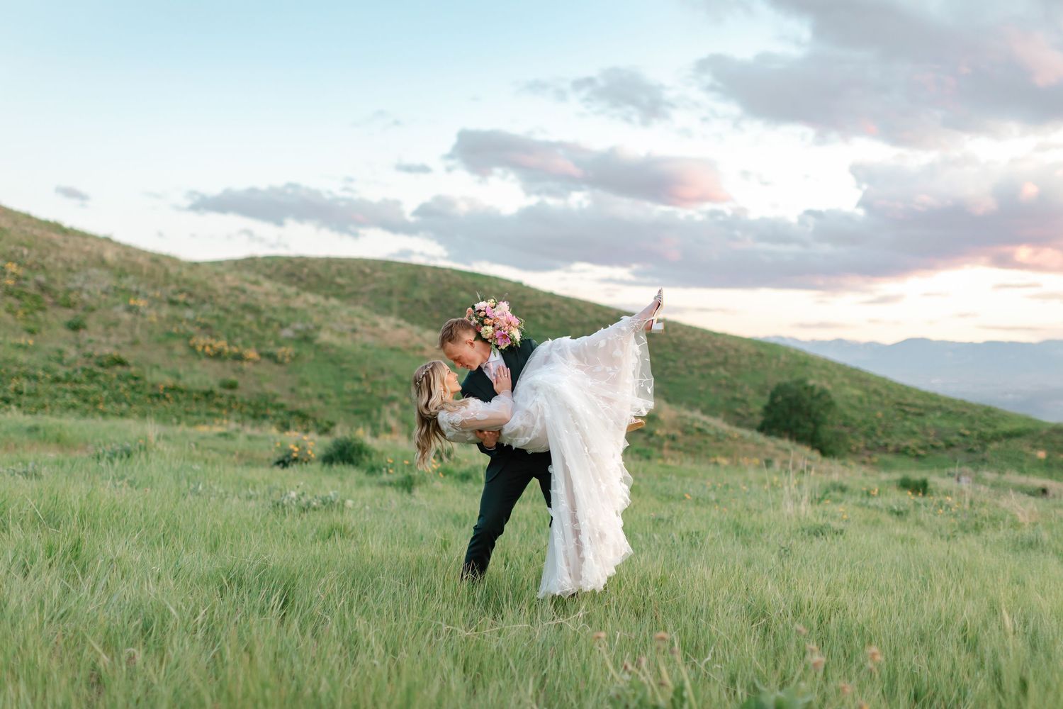 A romantic wedding couple shares a dip kiss in a scenic green meadow at sunset.