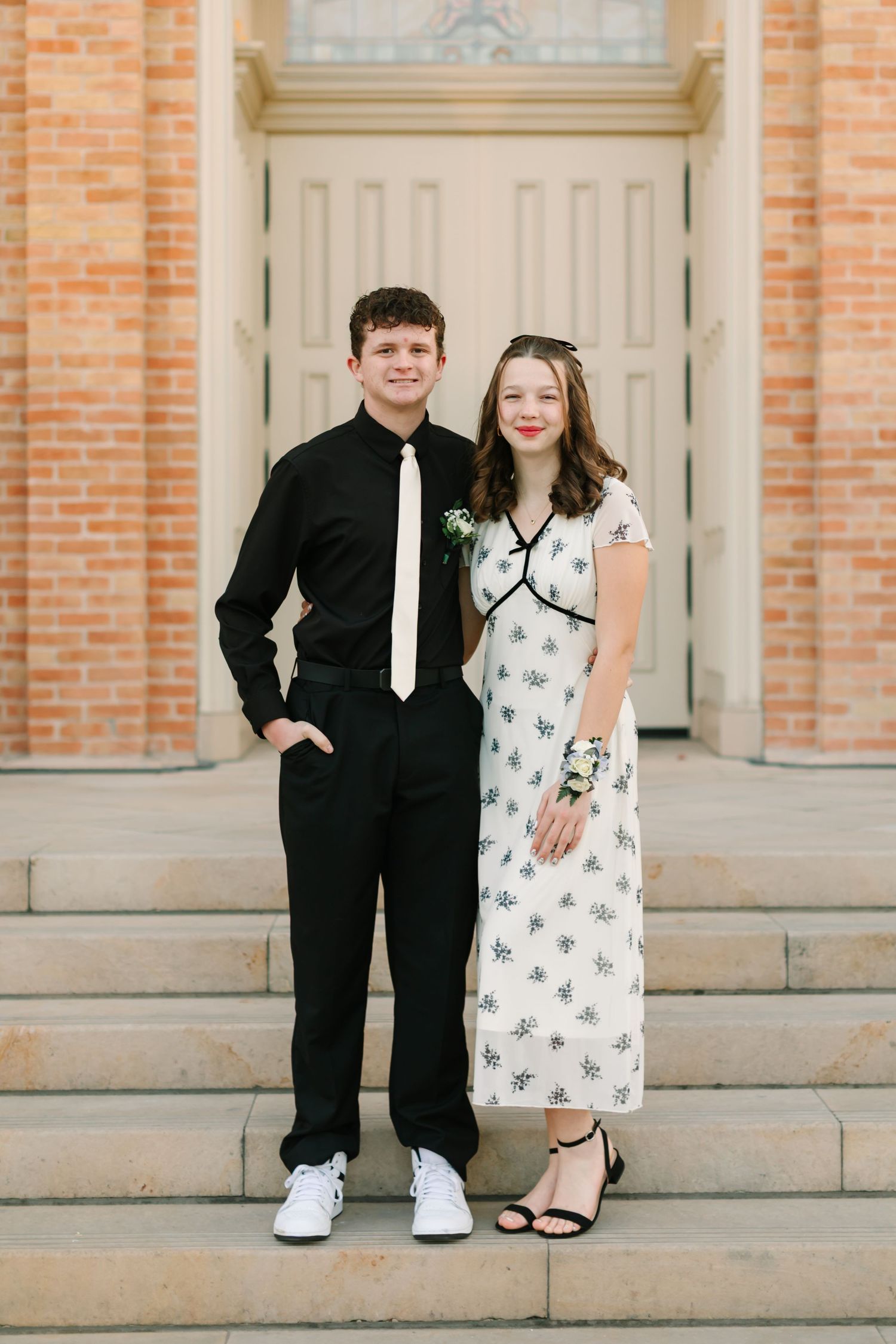 A couple in formal attire poses on stone steps in front of a brick building with white columns.