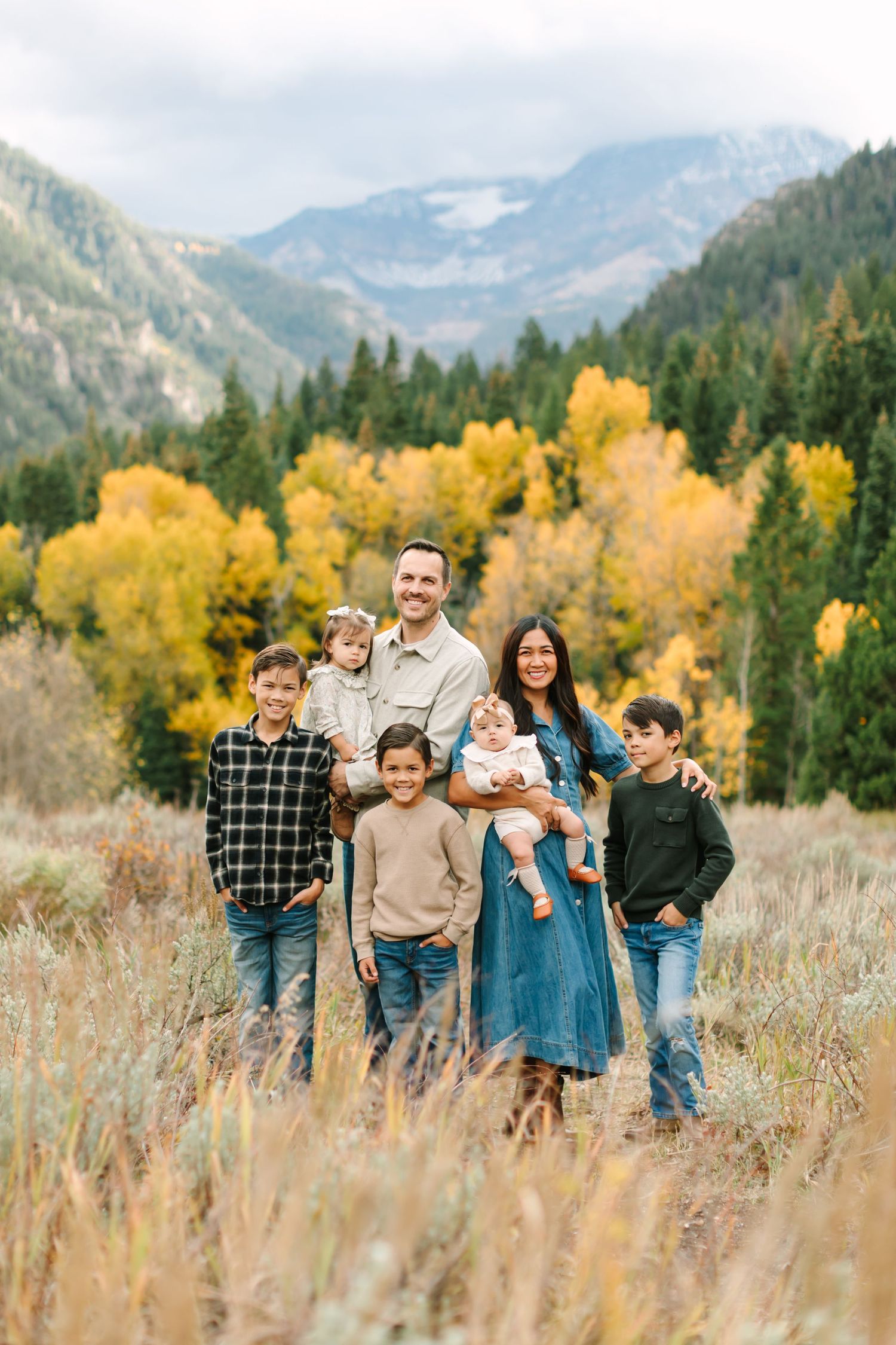 A family stands together in a field with golden autumn trees and mountains in the background.