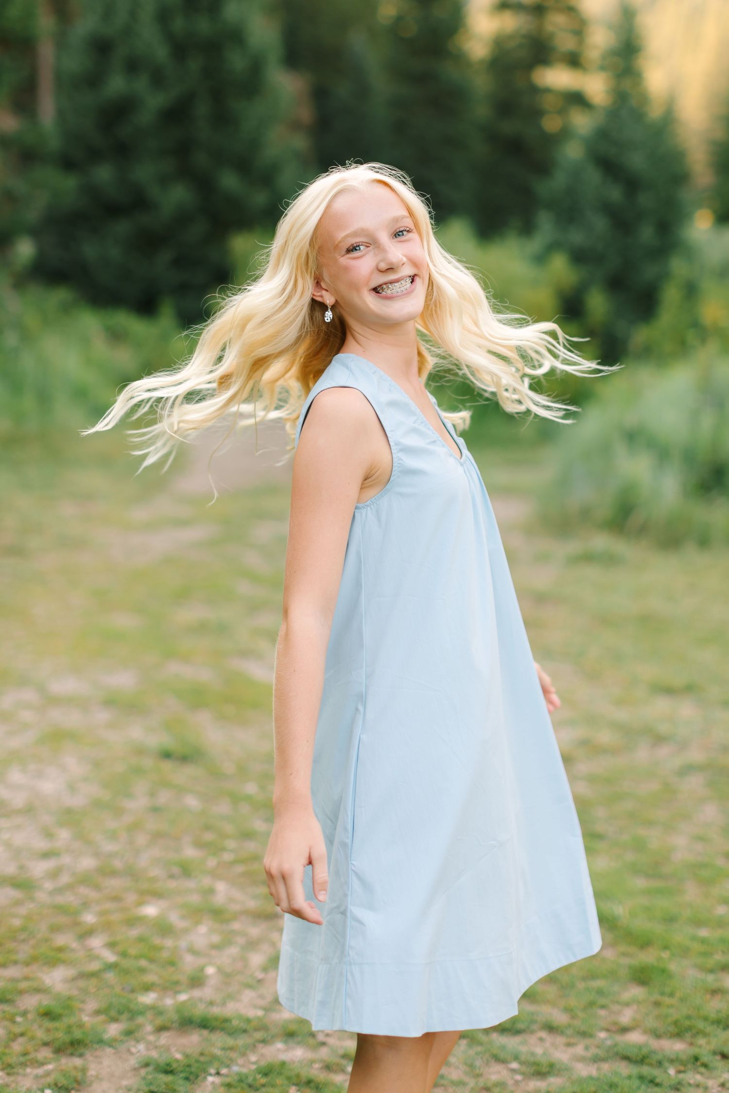 Smiling blonde girl in a light blue sleeveless dress twirling outdoors with green trees in the background.