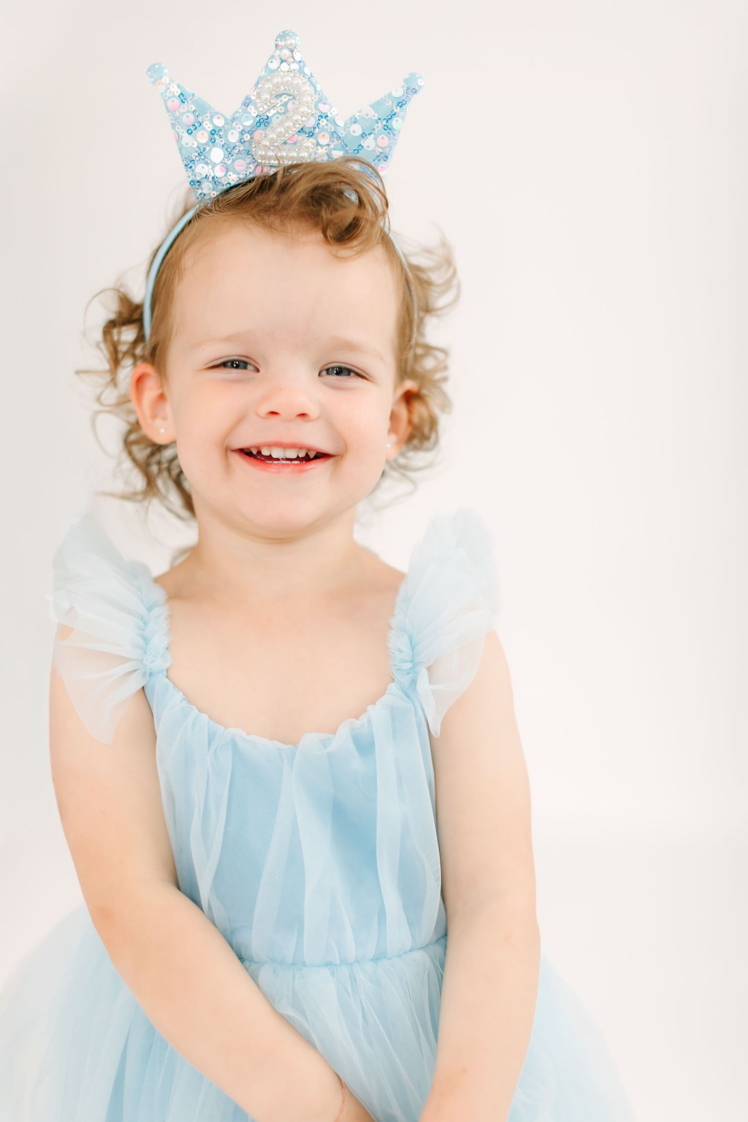 A young girl with curly hair wearing a blue dress and a sparkly crown, smiling happily.
