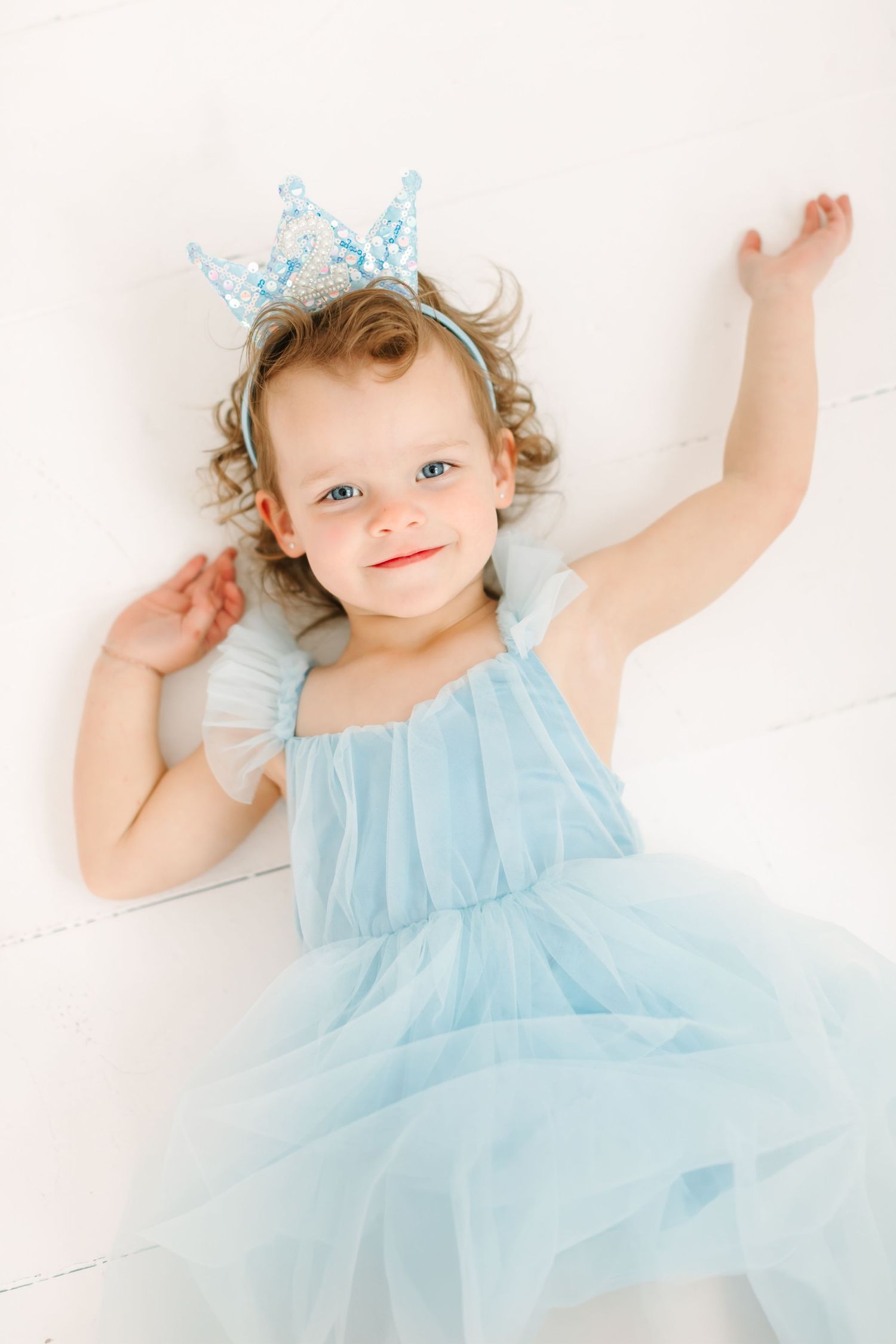 A young girl wearing a blue tulle dress and a crown-like headpiece smiles and raises her arms up.