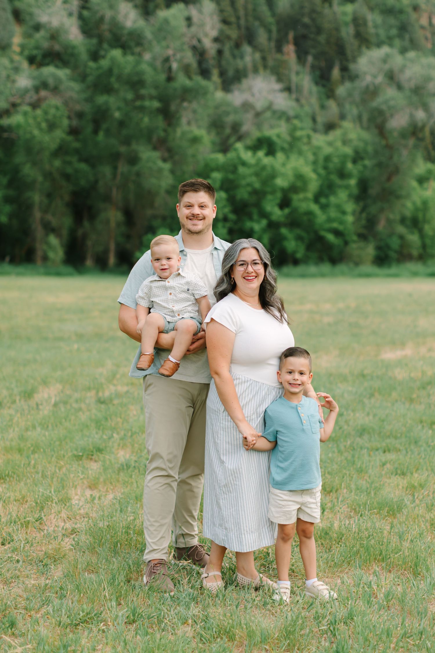 Family standing on green grass with green trees behind them at their family mini photography session at Jolly's Ranch in Springville, Utah.