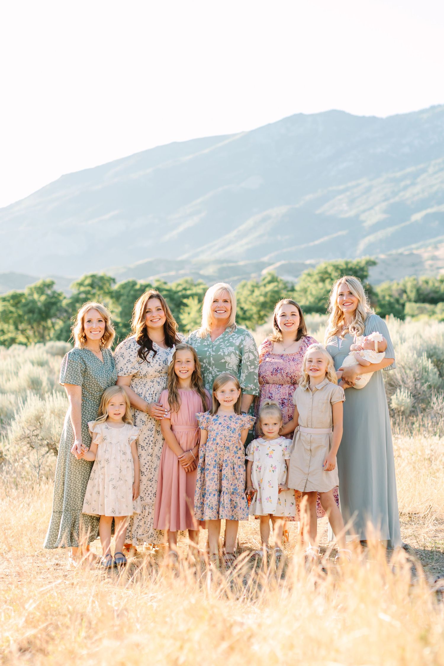 Group in coordinating floral and pastel dresses pose together in a golden field with mountains in background.