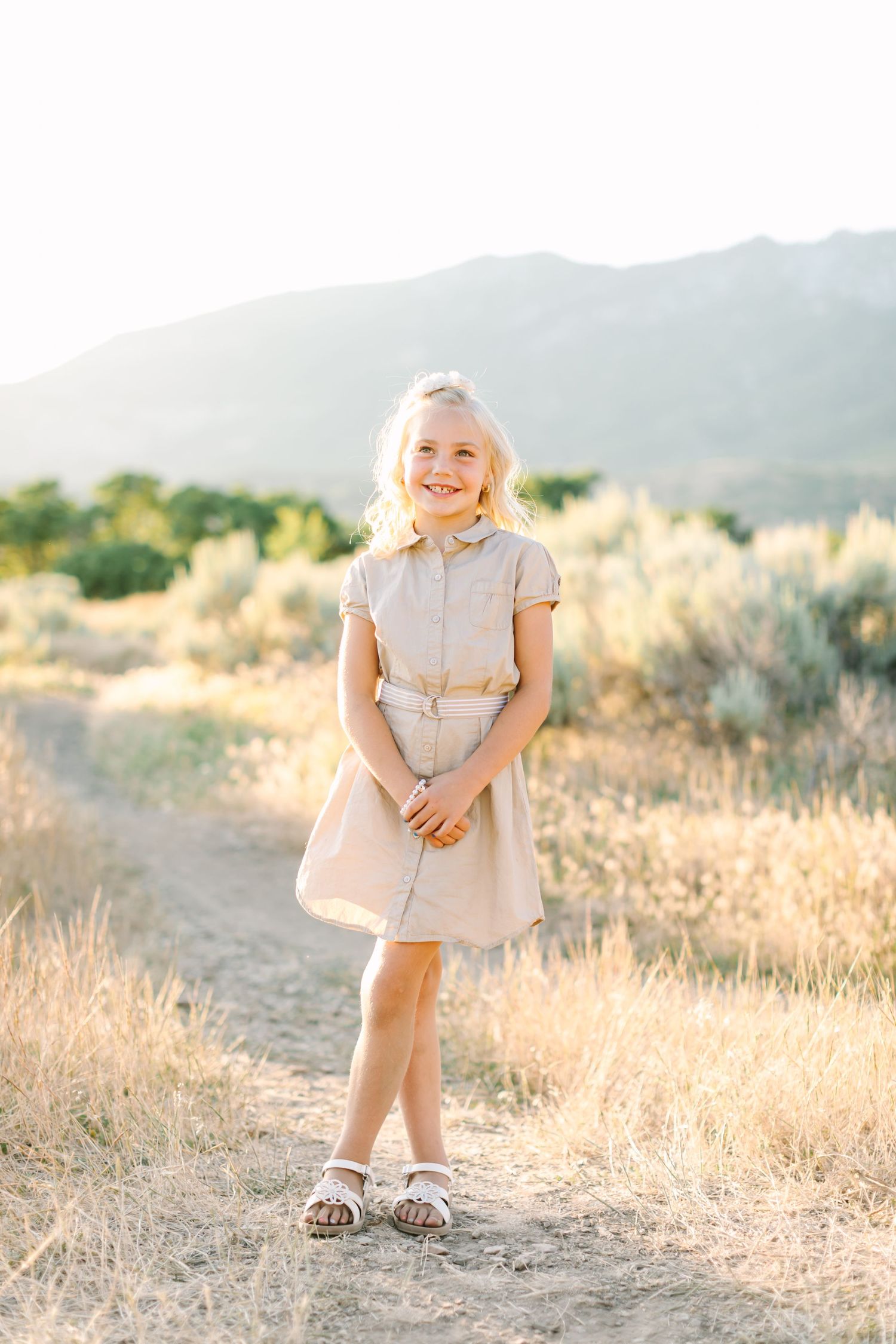 A person in a light beige dress stands in a sunlit field of golden grass with mountains in the background.