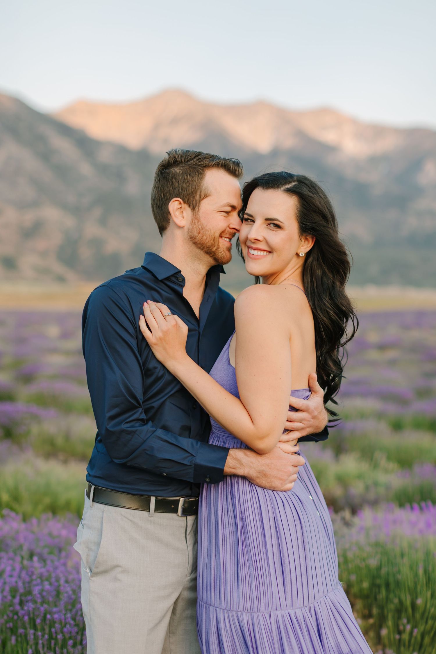 A couple embraces in a field of purple wildflowers with mountains in the background during sunset.