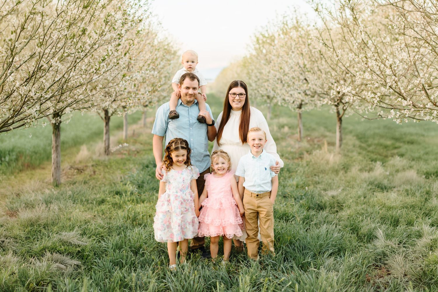 Family of six posing outdoors in a blooming orchard with white spring blossoms, parents with four young children smiling.