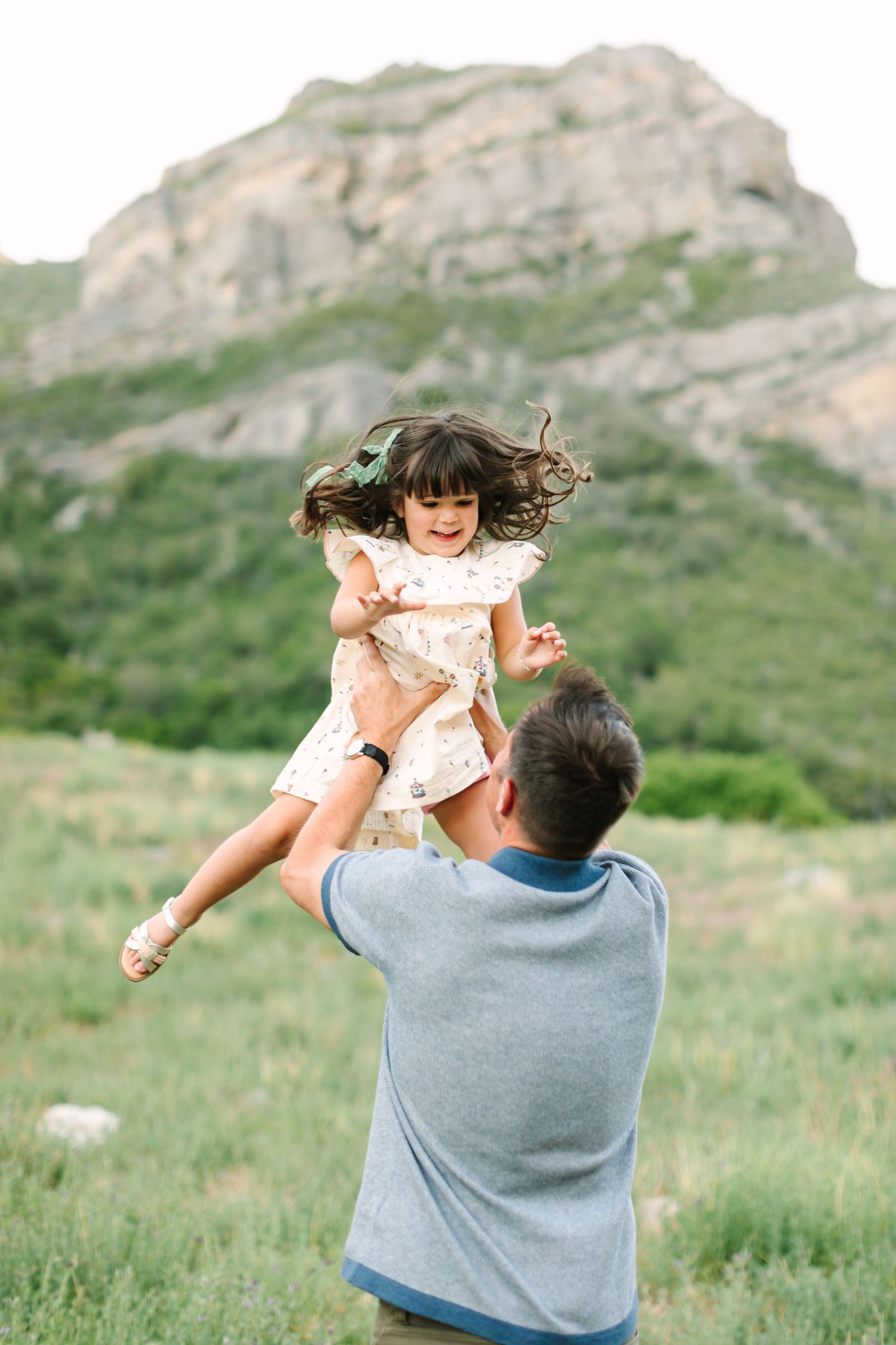 A joyful father lifts his laughing little girl in a floral dress outdoors with a rocky mountain backdrop.