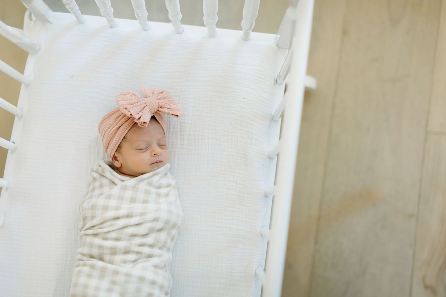 A sleeping infant wrapped in striped swaddle blanket rests in a white wooden crib.