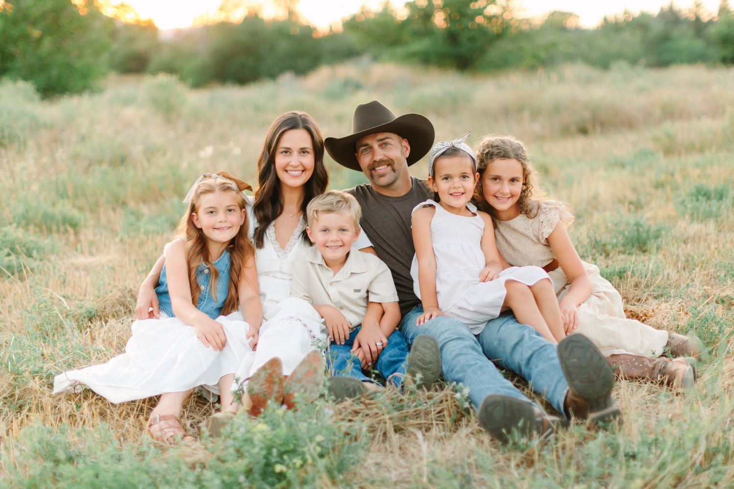 A family sits together in a grassy field at sunset wearing coordinated casual outfits in soft neutral colors.