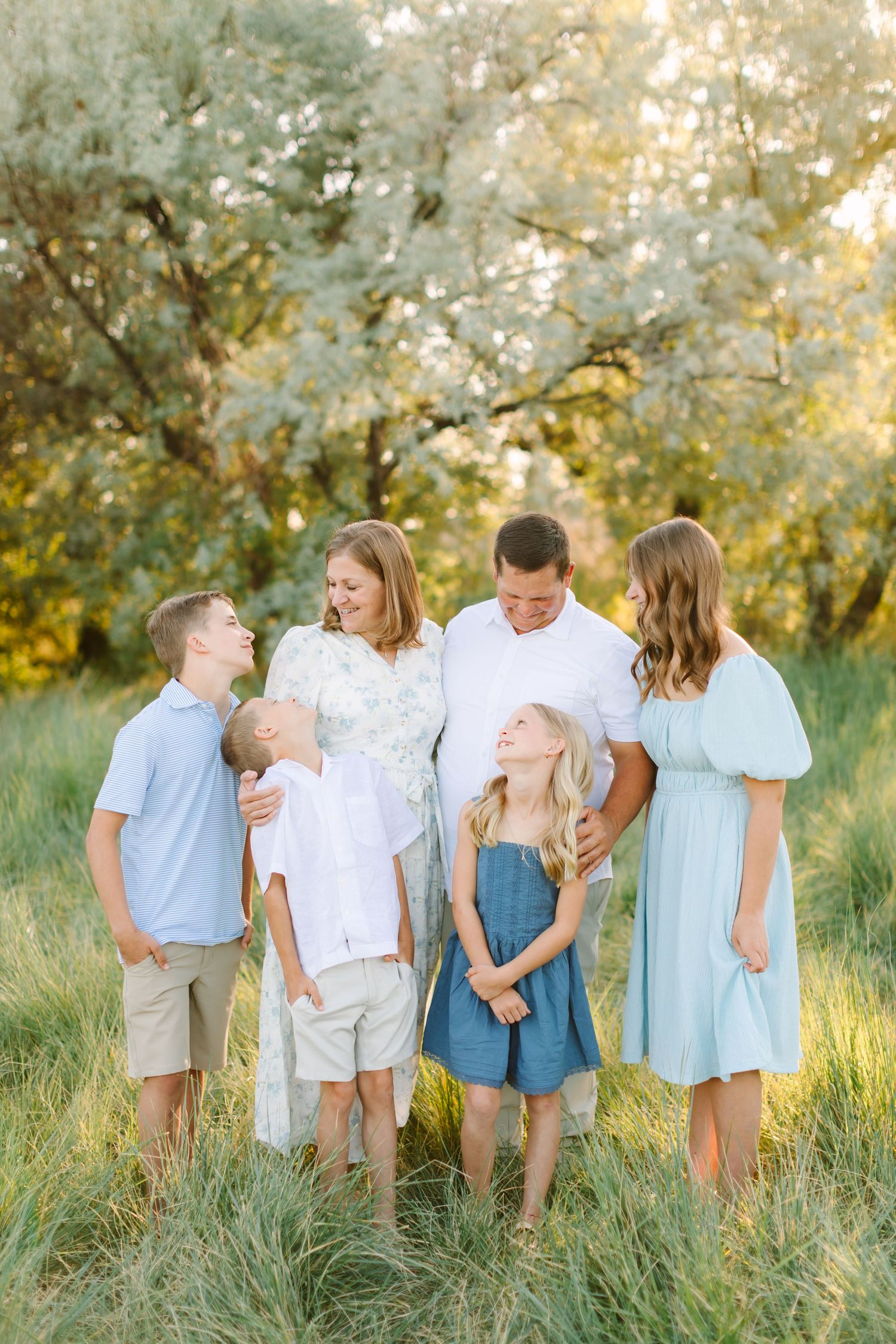 Family of six posing together in a sunny green meadow, smiling and laughing, dressed in light blue and white outfits.