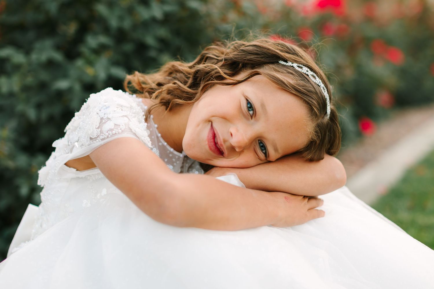 A bride in a white wedding dress poses gracefully in a garden setting with red flowers in the background.