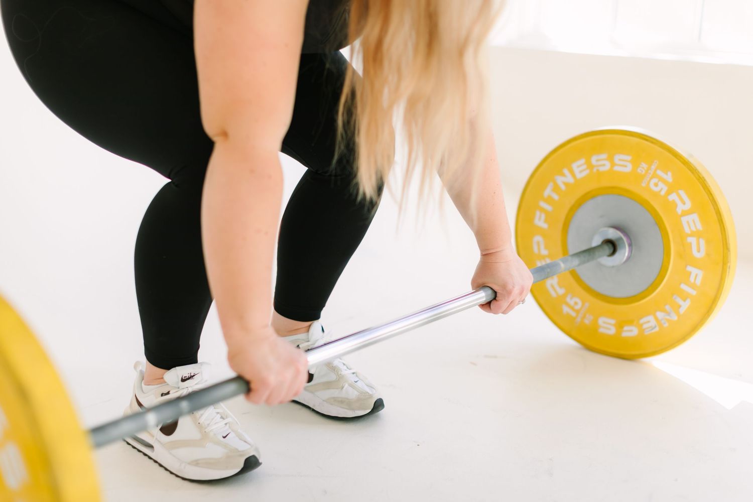 Person performing a deadlift exercise with yellow weight plates on a barbell in a bright indoor setting.