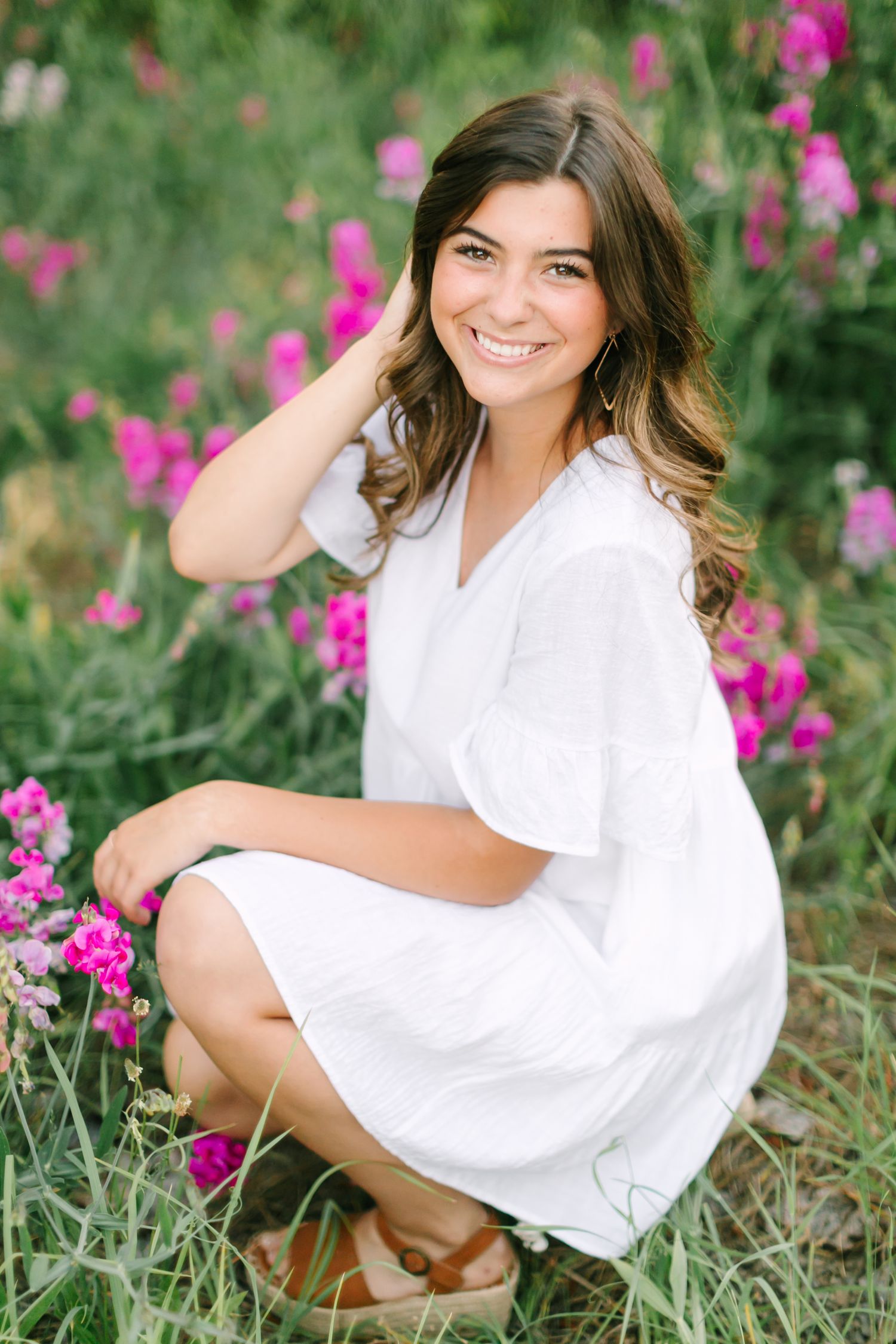 A person in a white dress sits in a field of vibrant pink wildflowers during a sunny outdoor photo session.