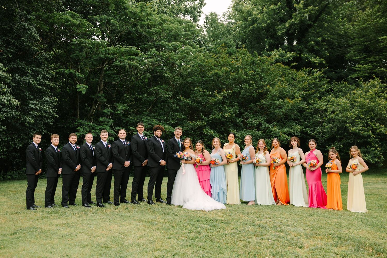 Wedding party lined up outdoors with bridesmaids in rainbow colored dresses and groomsmen in black suits.