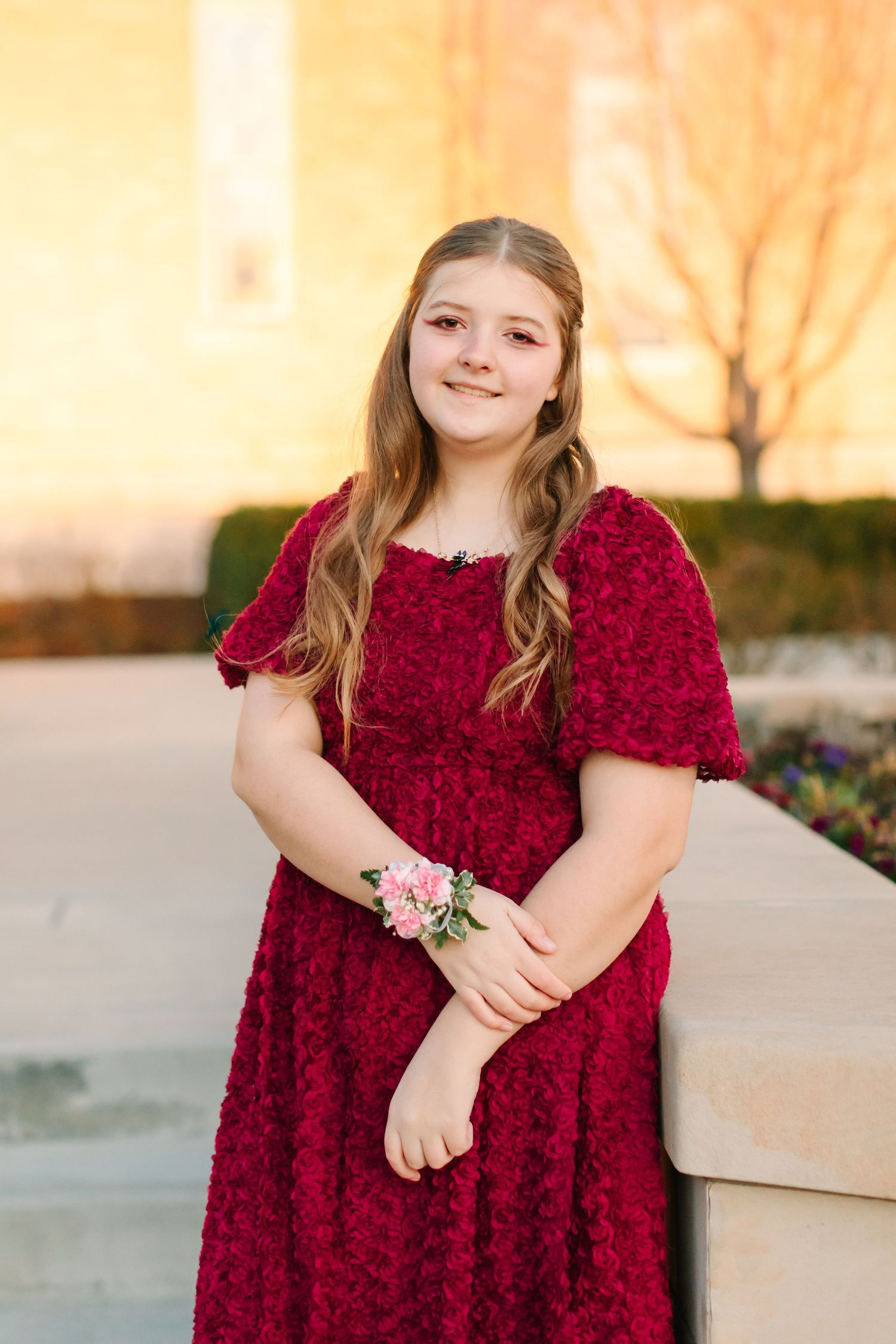 Young person in a burgundy velvet dress with floral wrist corsage poses outdoors at sunset.