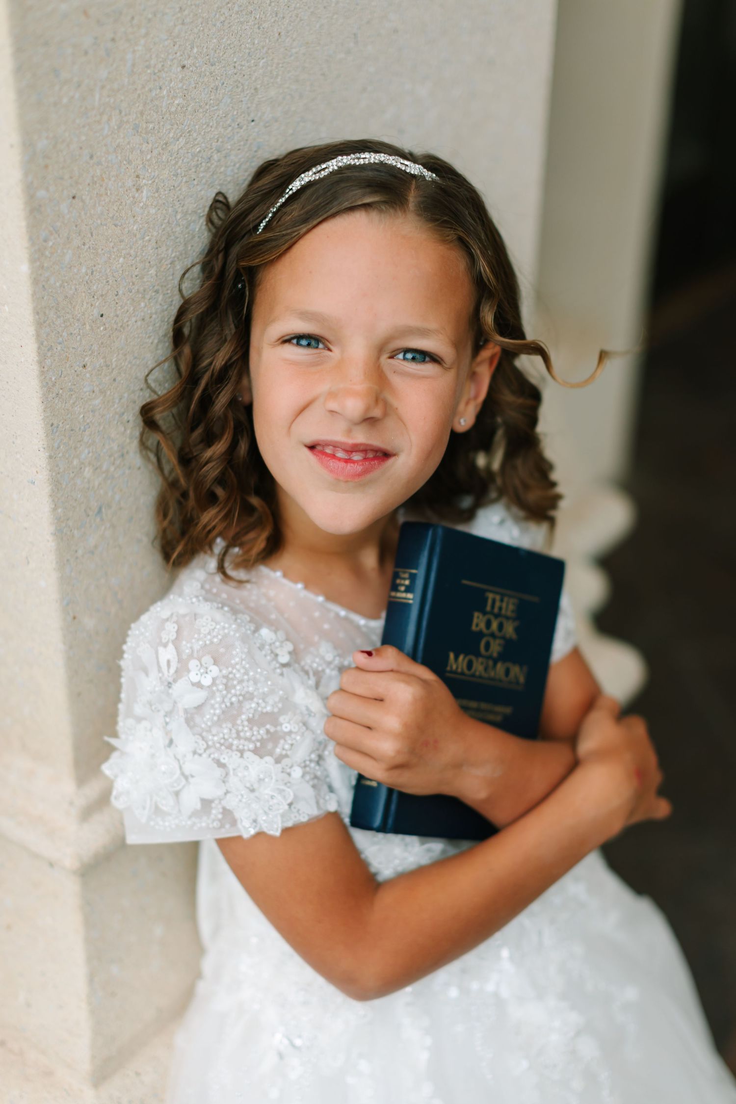 A child in a white lace dress holds a small blue book while smiling at the camera for her LDS Baptism in Payson, Utah in Utah County.