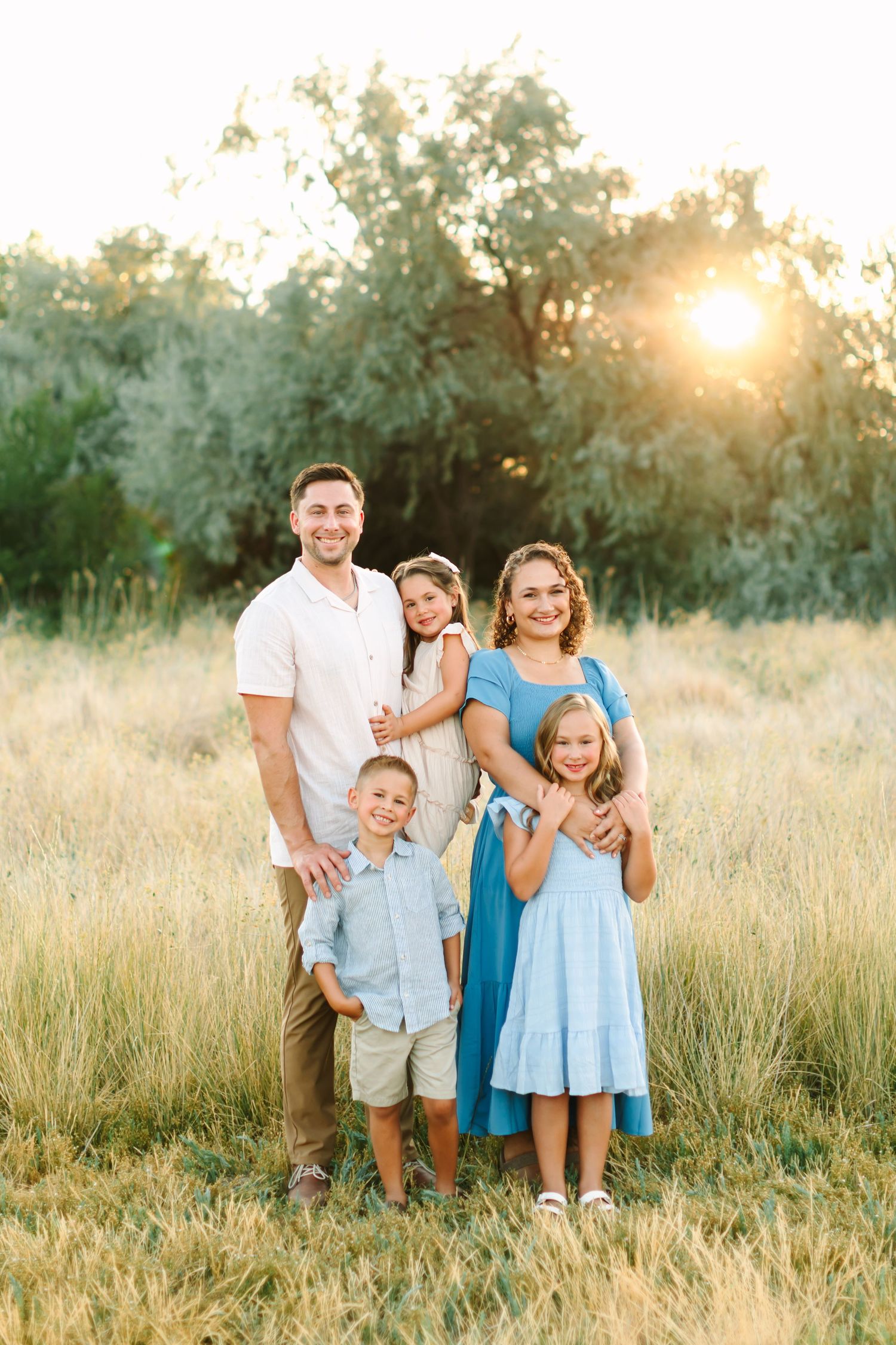 Family of four posing in golden field at sunset, parents holding children in blue and white outfits in Bountiful, Utah.