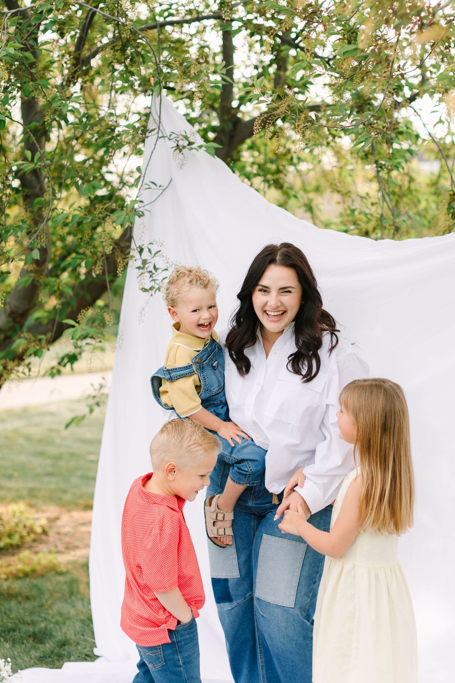 A smiling dark-haired woman in white shirt and jeans holds a toddler while two young children stand nearby outdoors.