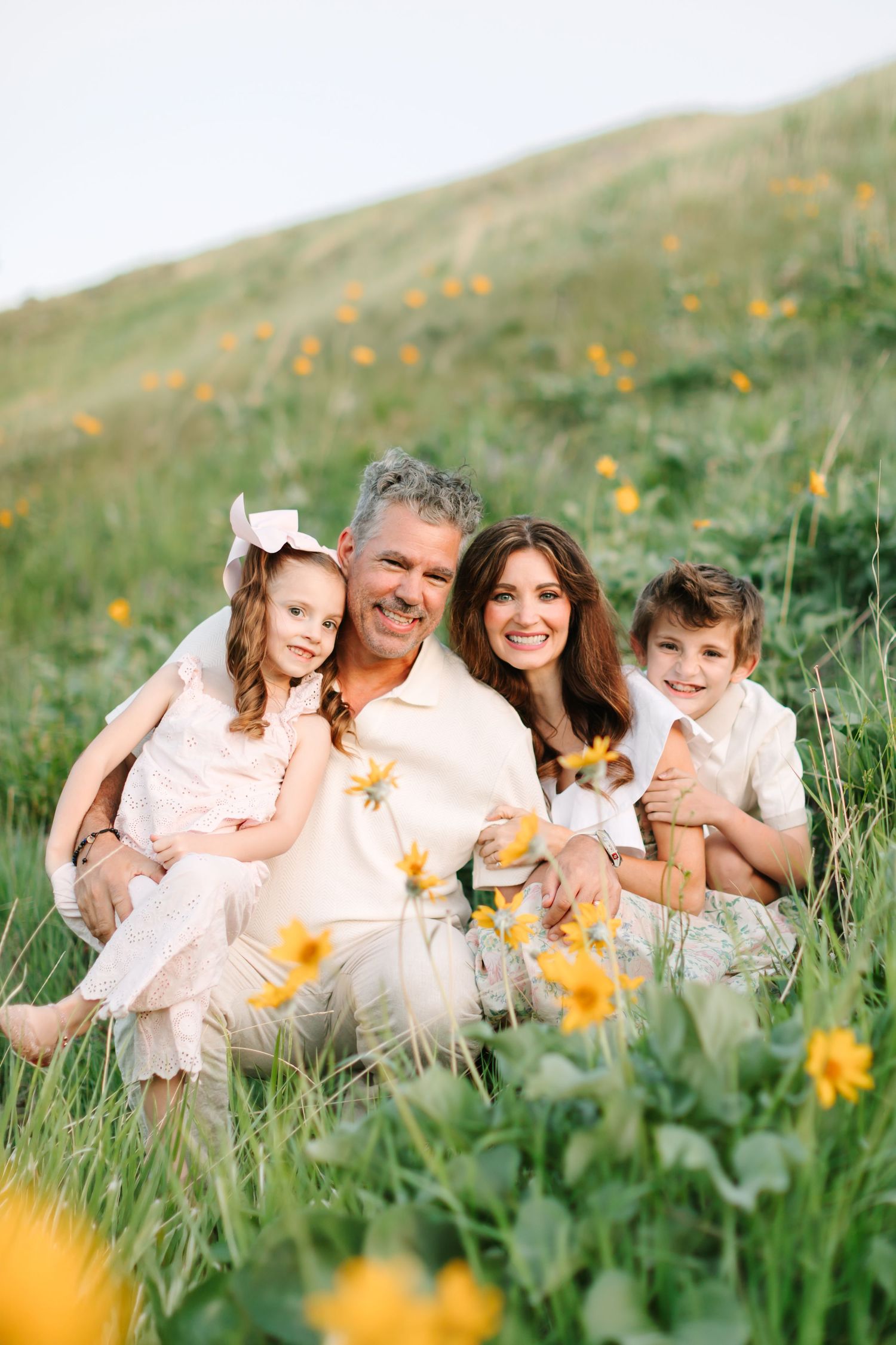 Happy family of four sitting in a field of yellow wildflowers, smiling outdoors on a sunny day.