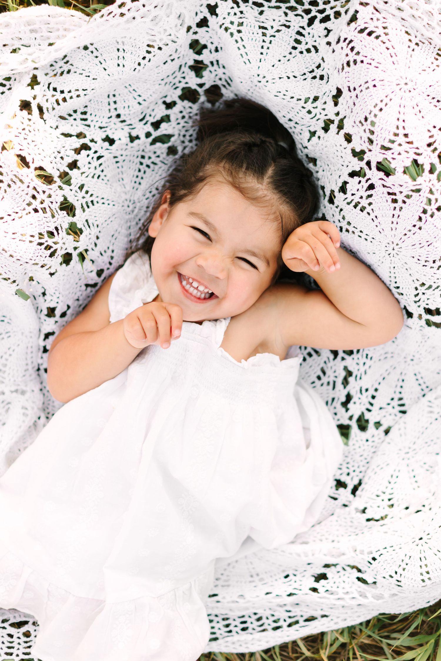 A joyful young girl in a white dress laughs while lying on a white lace blanket outdoors.
