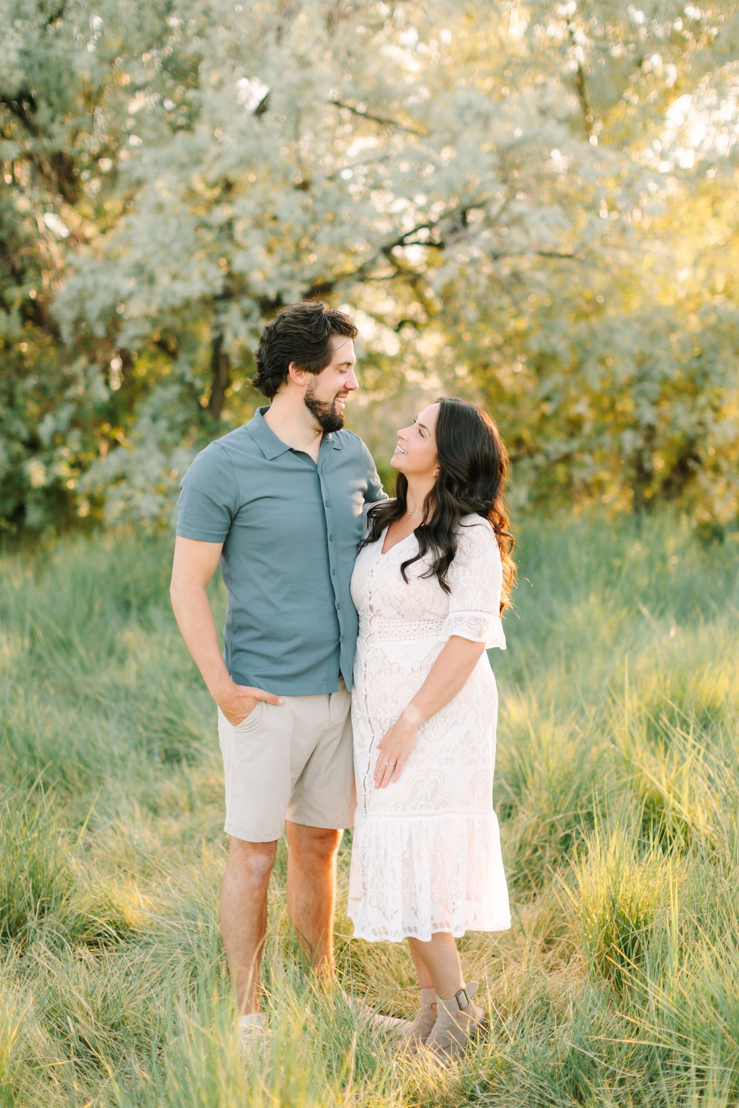 A couple gazes lovingly at each other in a sunlit meadow, man in blue polo and shorts, woman in white lace dress.