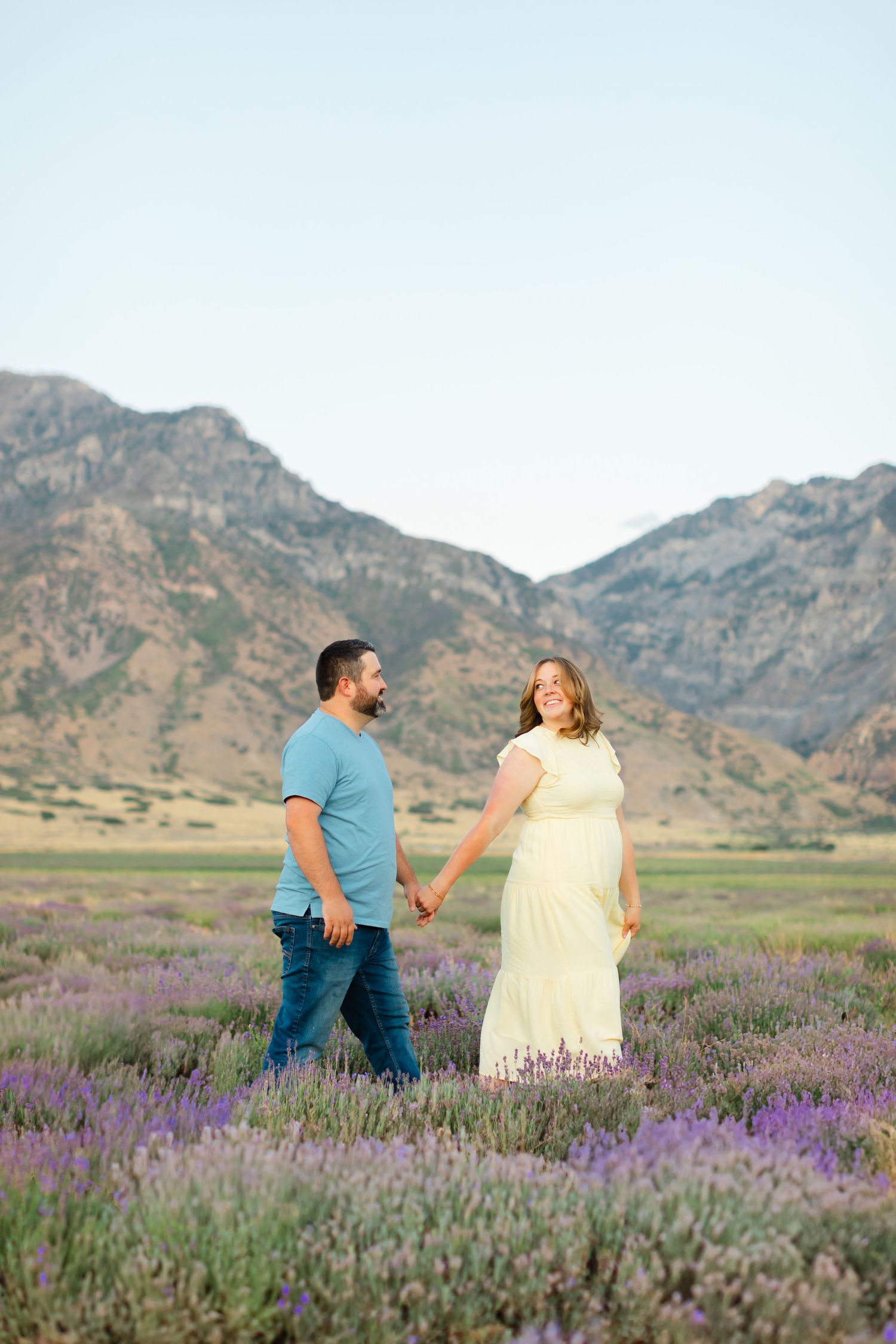 A couple stands together in a blooming purple lavender field with rugged mountains rising in the background at sunset.